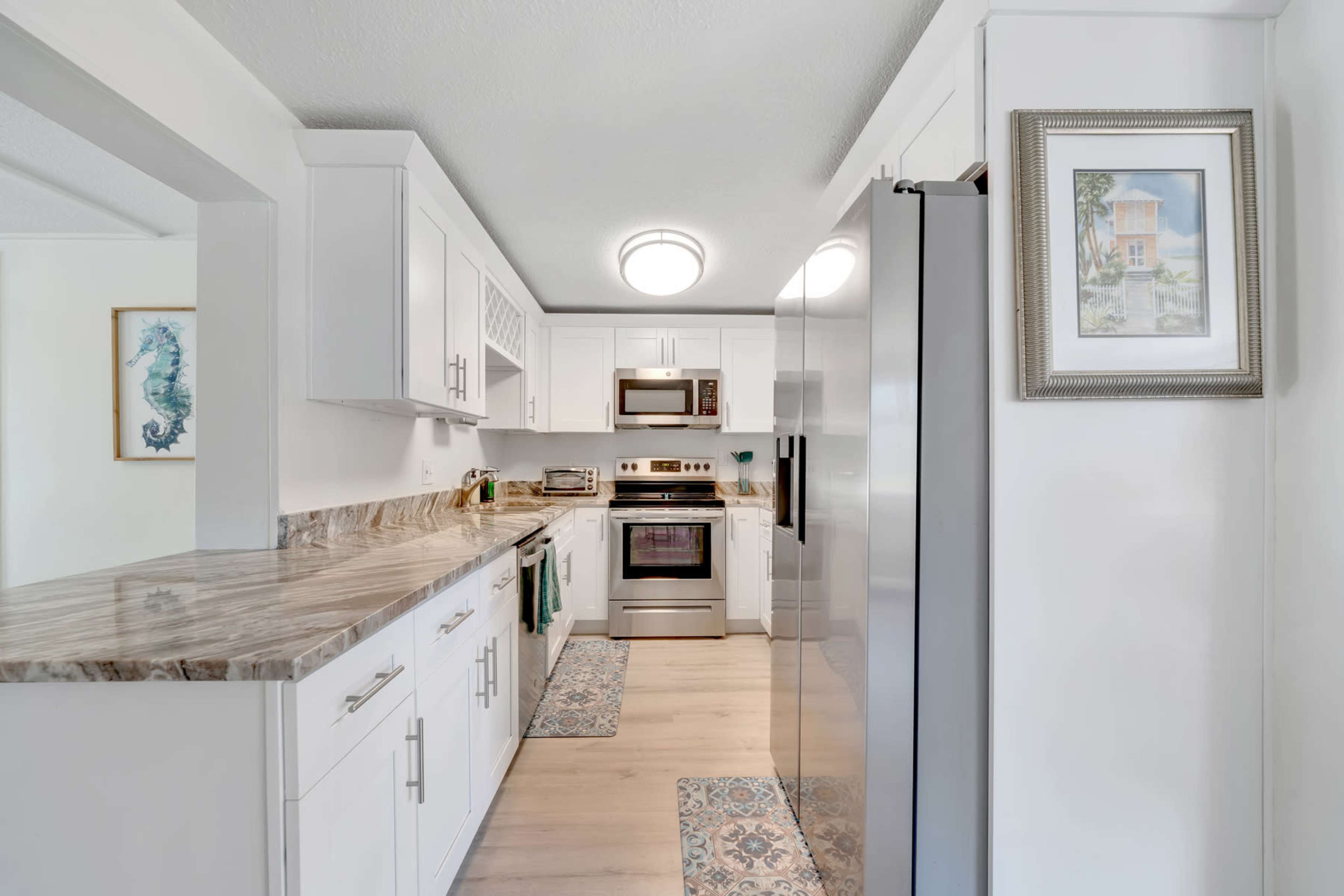 A well-lit kitchen features white cabinetry, stainless steel appliances, and a marble countertop with decorative rugs on a wooden floor.