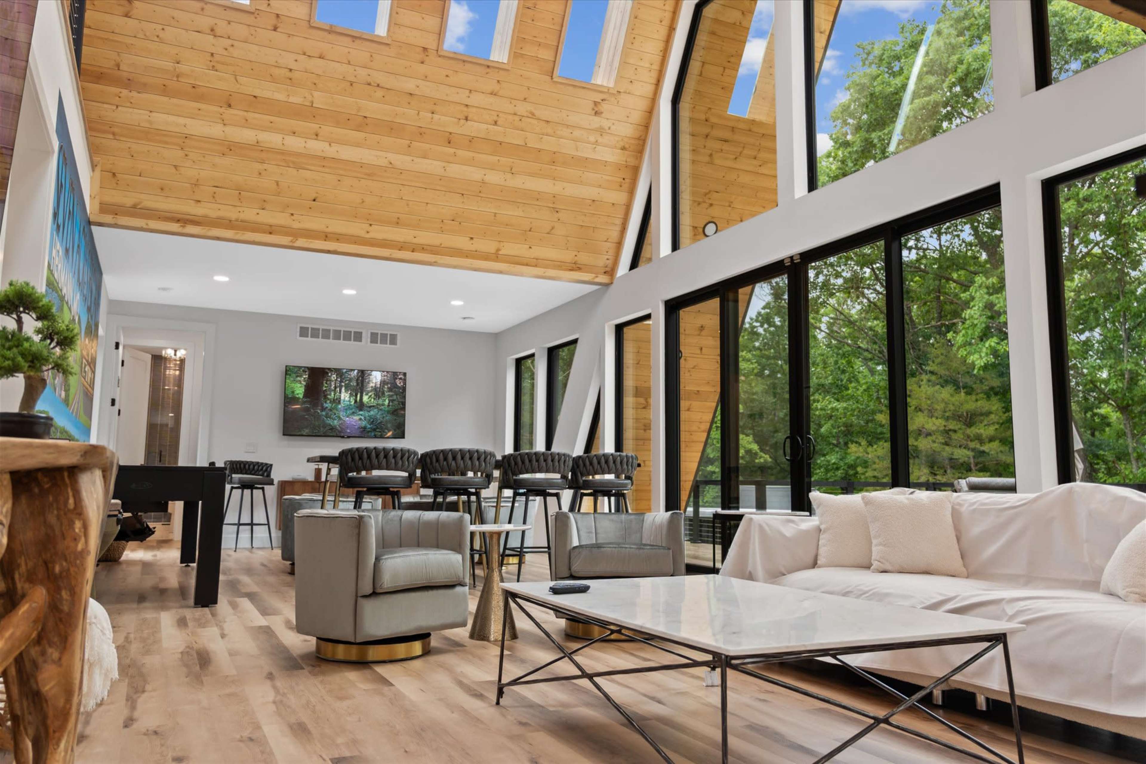 The image shows a modern living space with a high wooden ceiling, large windows, a white sofa, and black chairs arranged around a dining table.