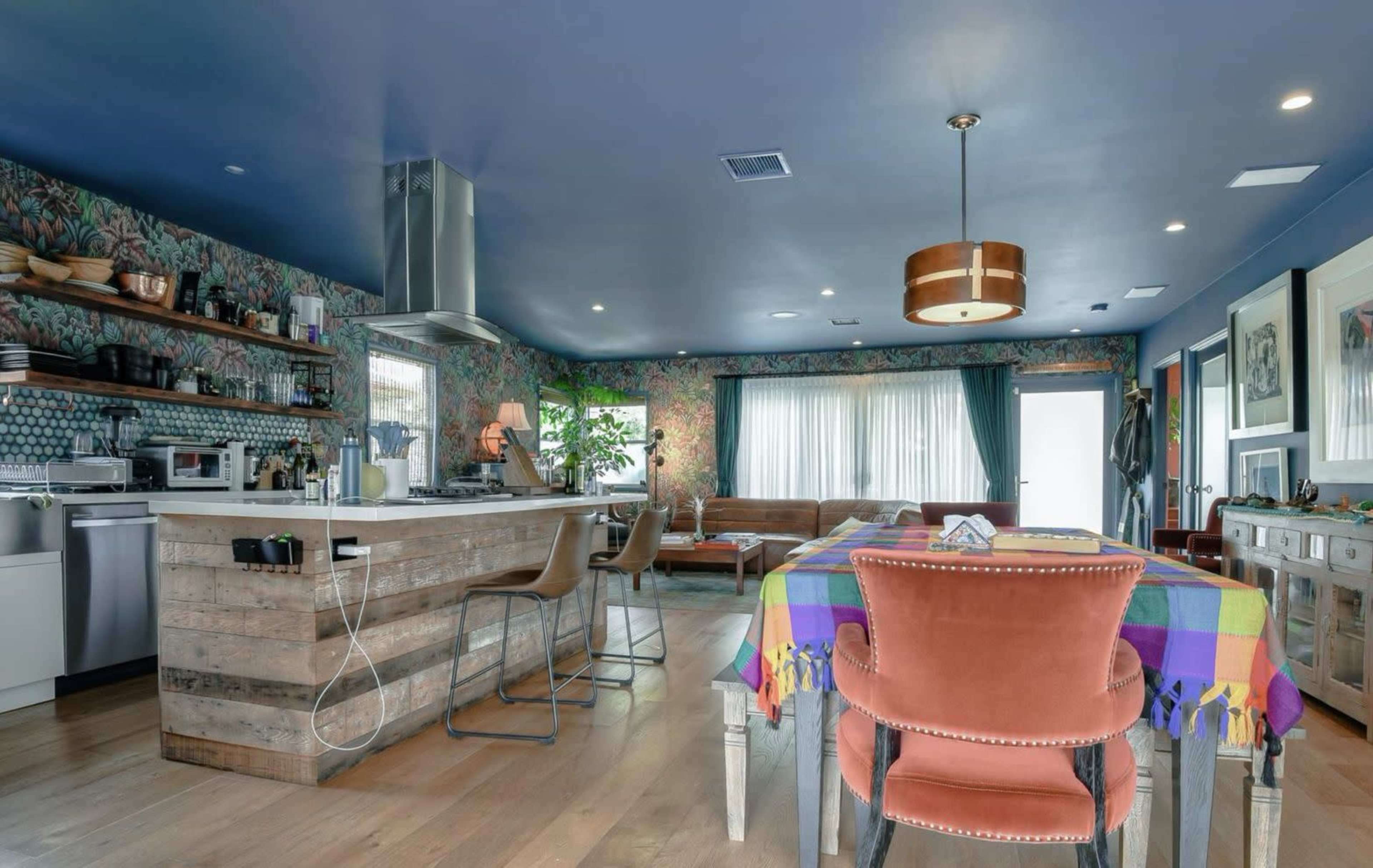 A modern kitchen and dining area featuring a large wooden table with colorful tablecloth, surrounded by chairs, and a patterned wallpaper accenting the walls.