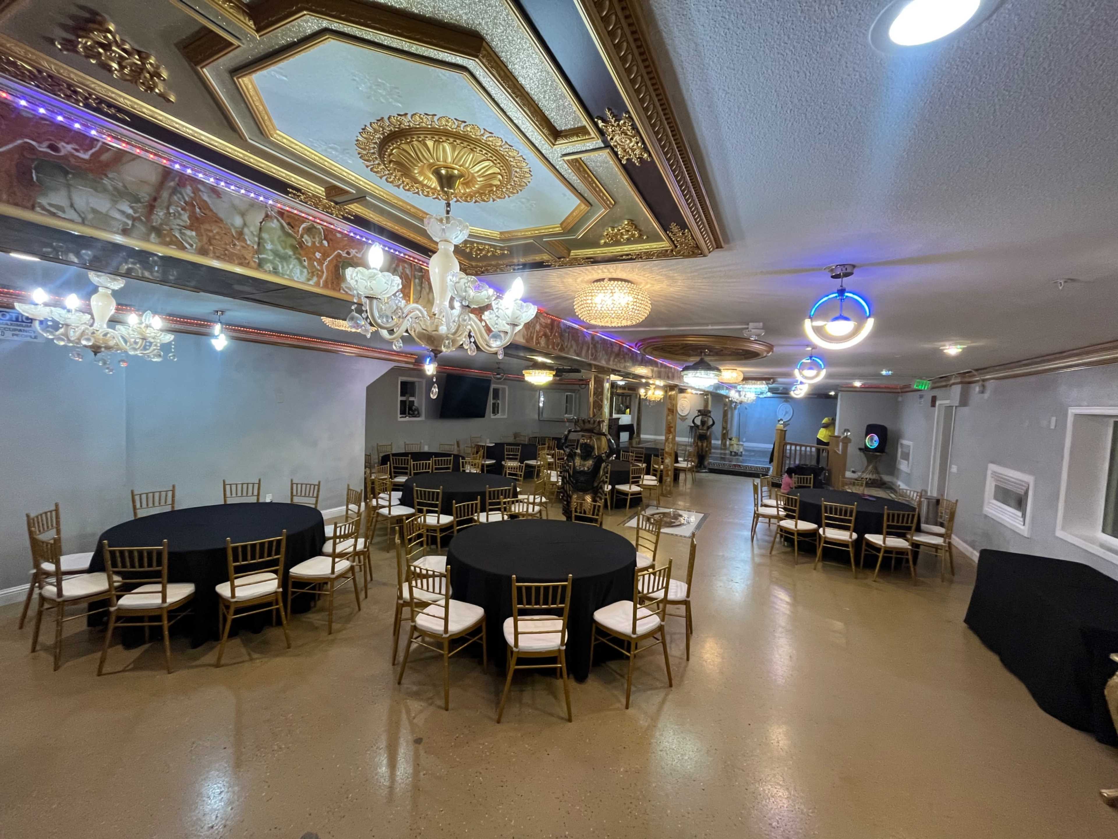 The image shows a spacious banquet room with gold-accented decor, featuring several round tables covered with black tablecloths and multiple chandeliers hanging from the ceiling.