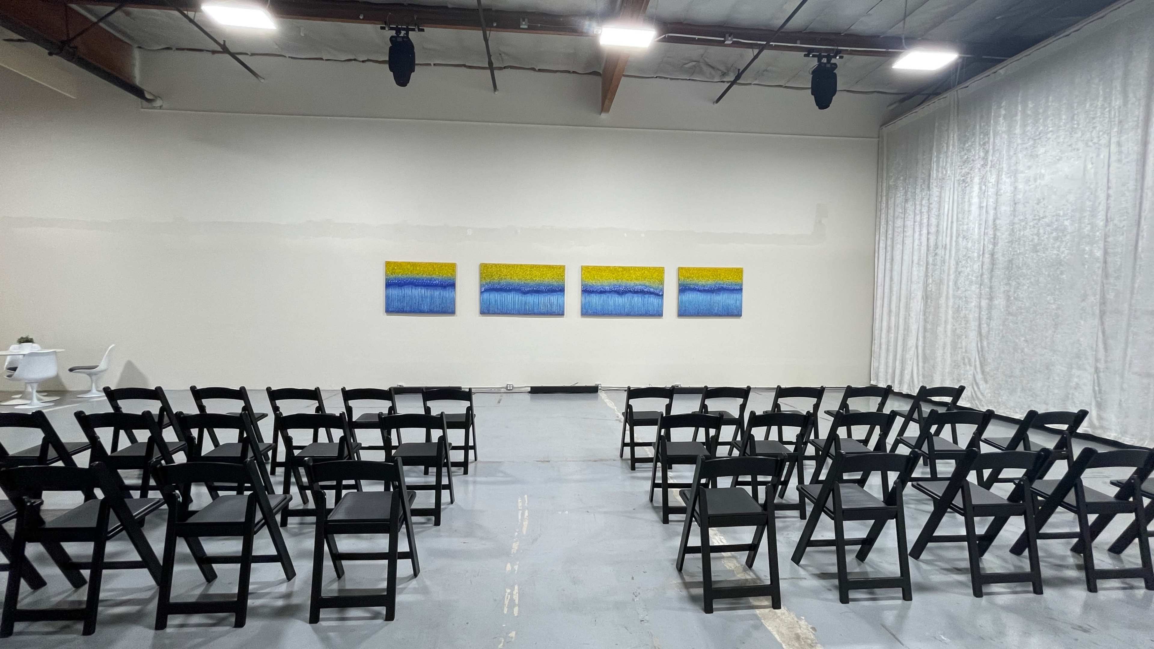 A row of black chairs facing a wall with four abstract paintings, set in a spacious, unfinished indoor venue.