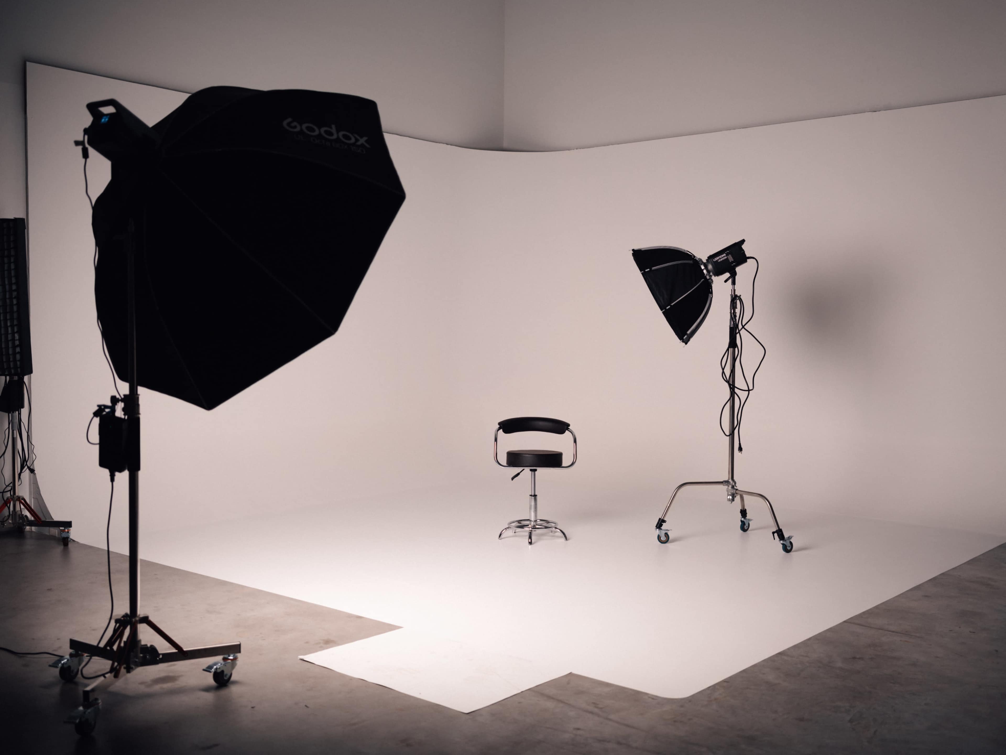 The image shows a photography studio with a white backdrop, a black stool in the center, and two large softbox lights positioned at different angles.