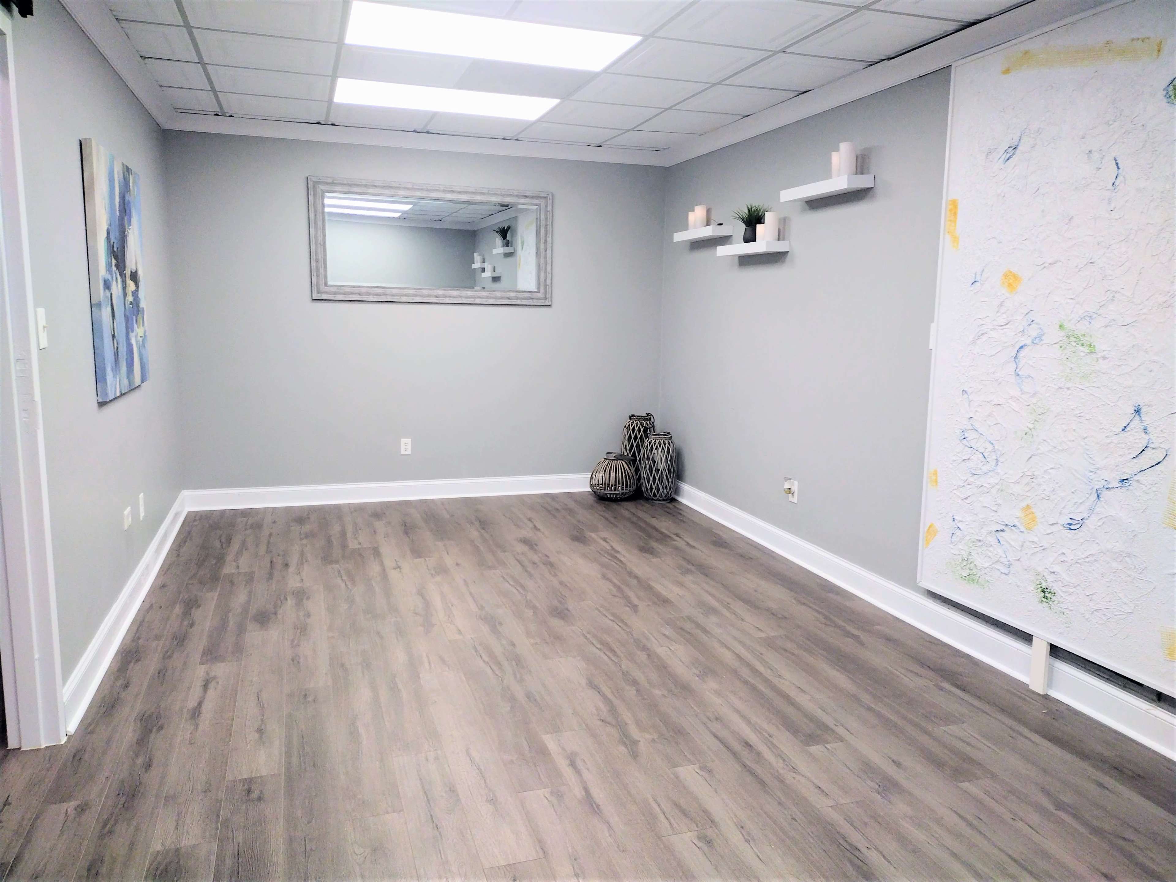 A spacious, empty room with gray walls, wooden flooring, a large mirror, and minimalist shelves displaying decorative items.