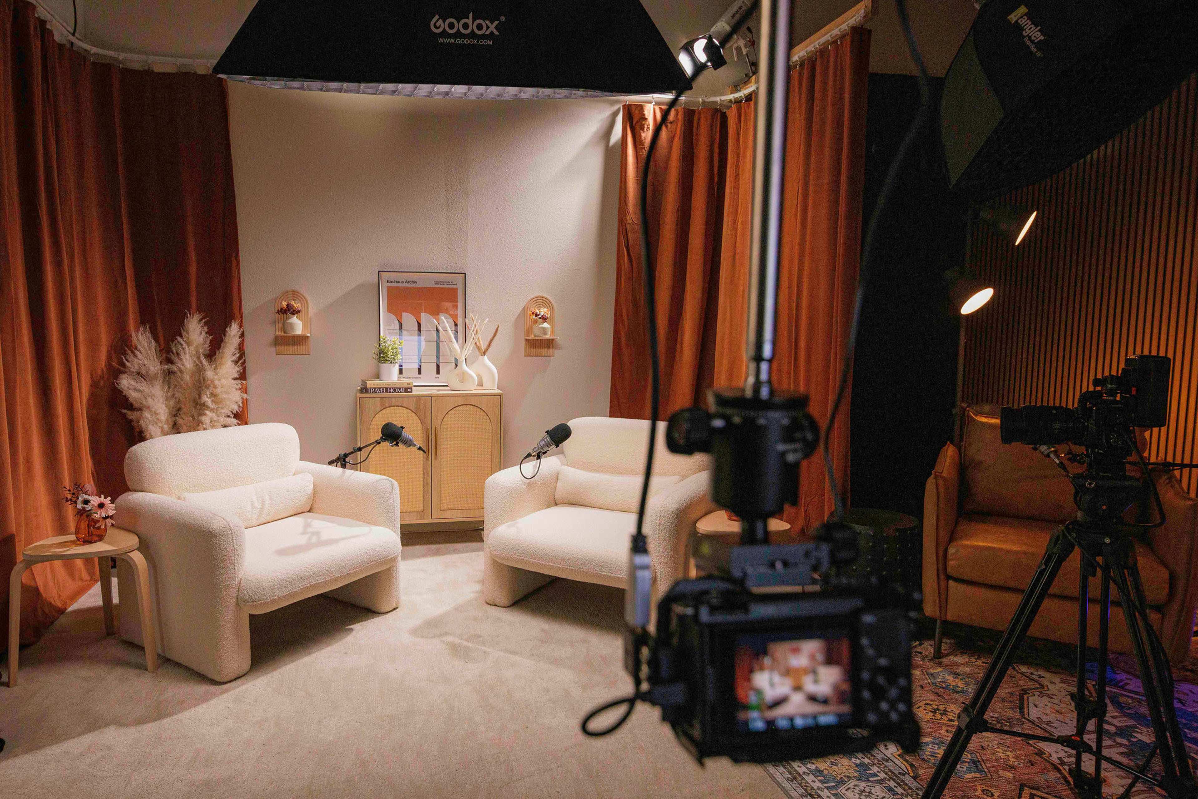 A well-lit studio setup featuring two white armchairs and various recording equipment, including cameras and microphones.