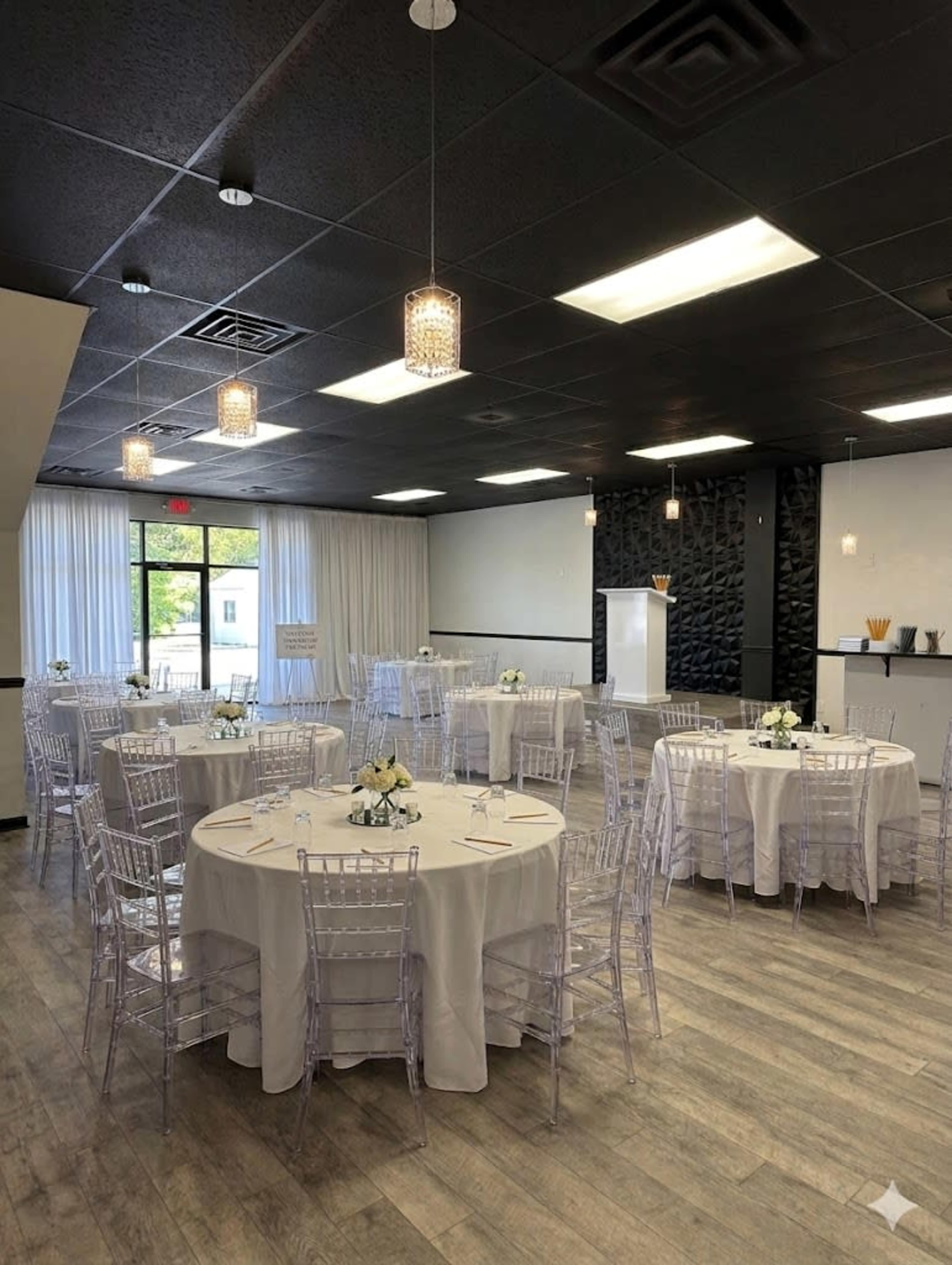 The image shows a modern event space with round tables set for a gathering, featuring white tablecloths and clear chiavari chairs, along with pendant lighting and a sleek bar area.