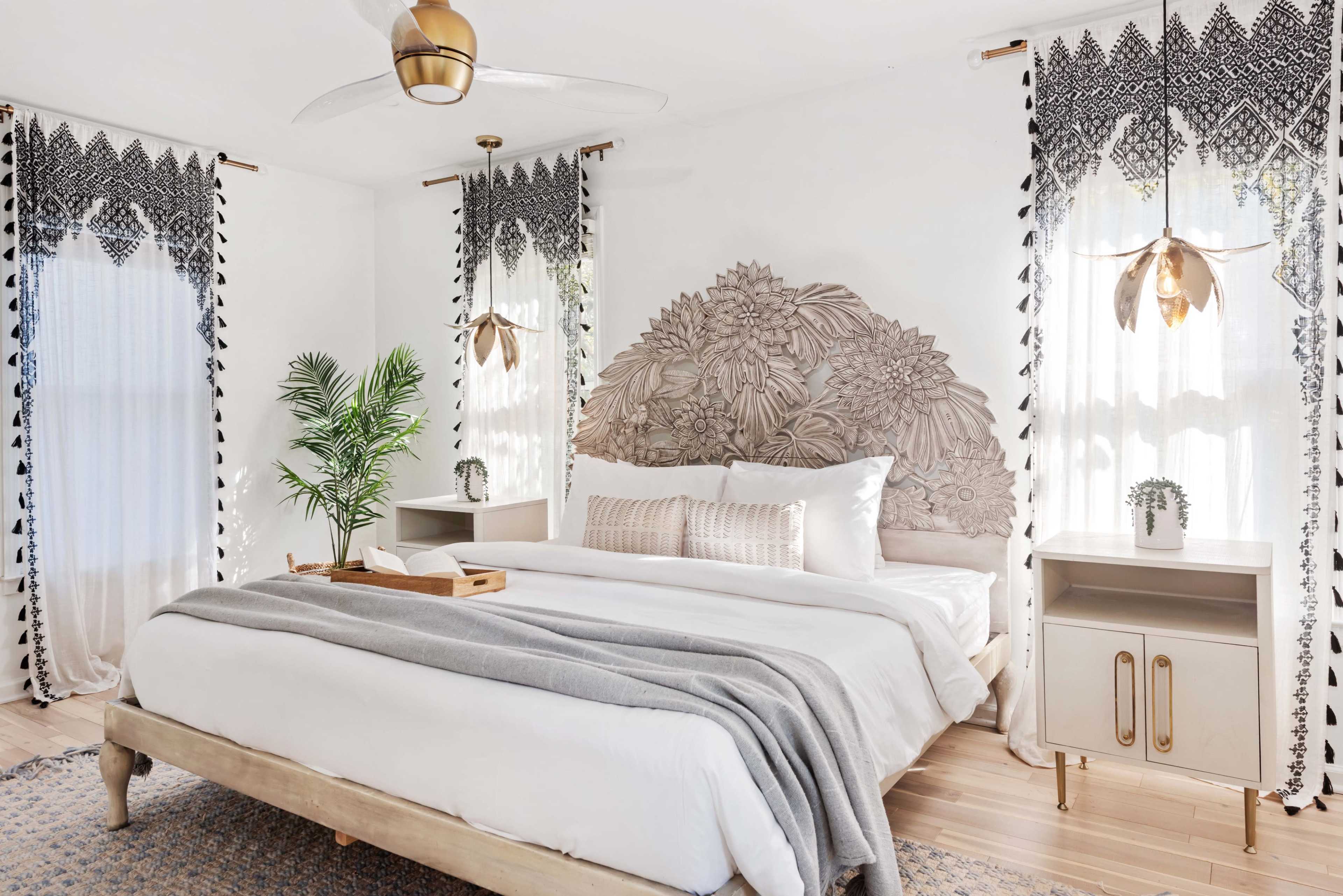 The image shows a bright bedroom featuring a large bed with a decorative headboard, flanked by two bedside tables and adorned with elegant curtains and a potted plant.