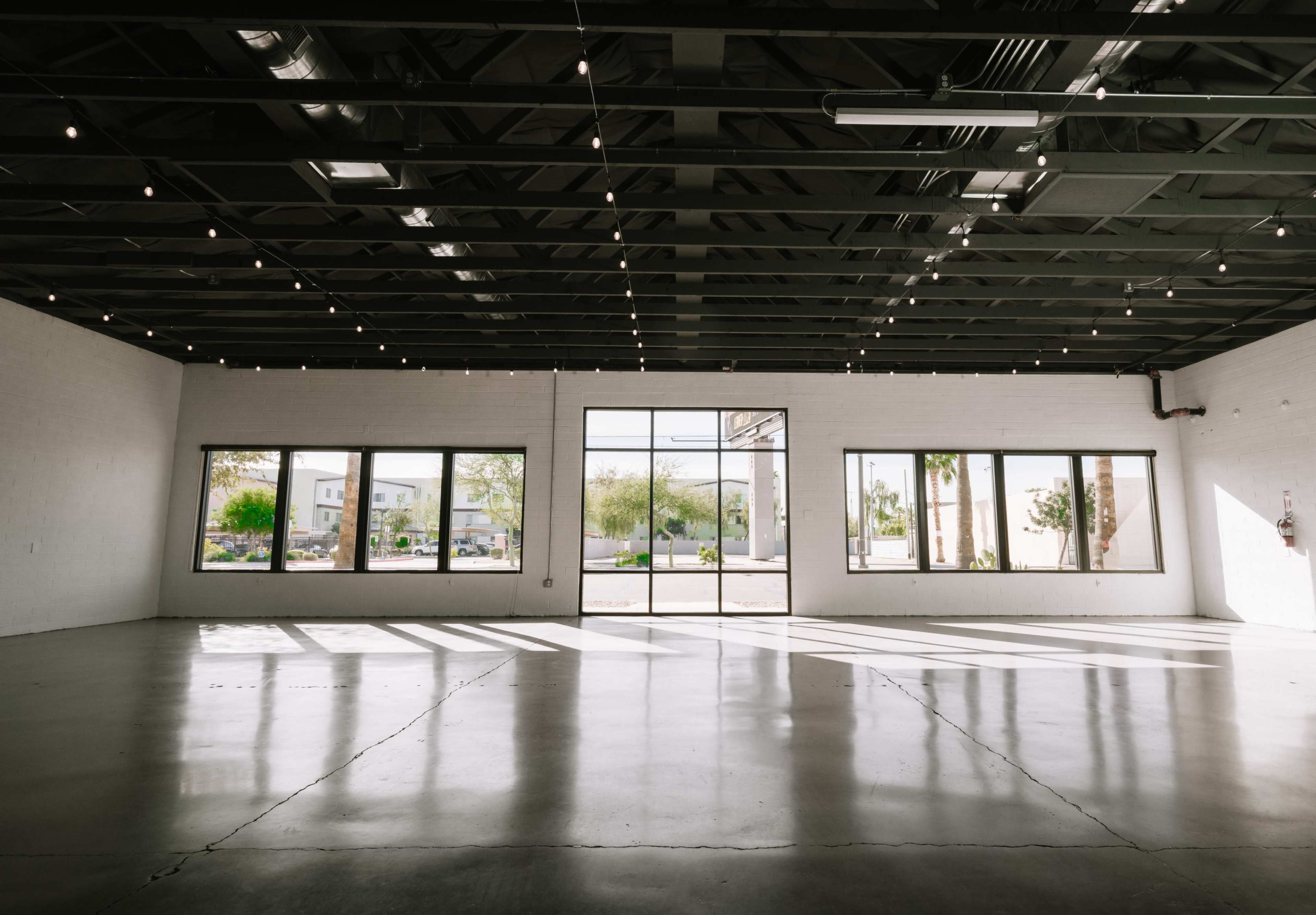 The image shows a spacious, empty room with polished concrete floors, large windows, and overhead string lights illuminating the area.