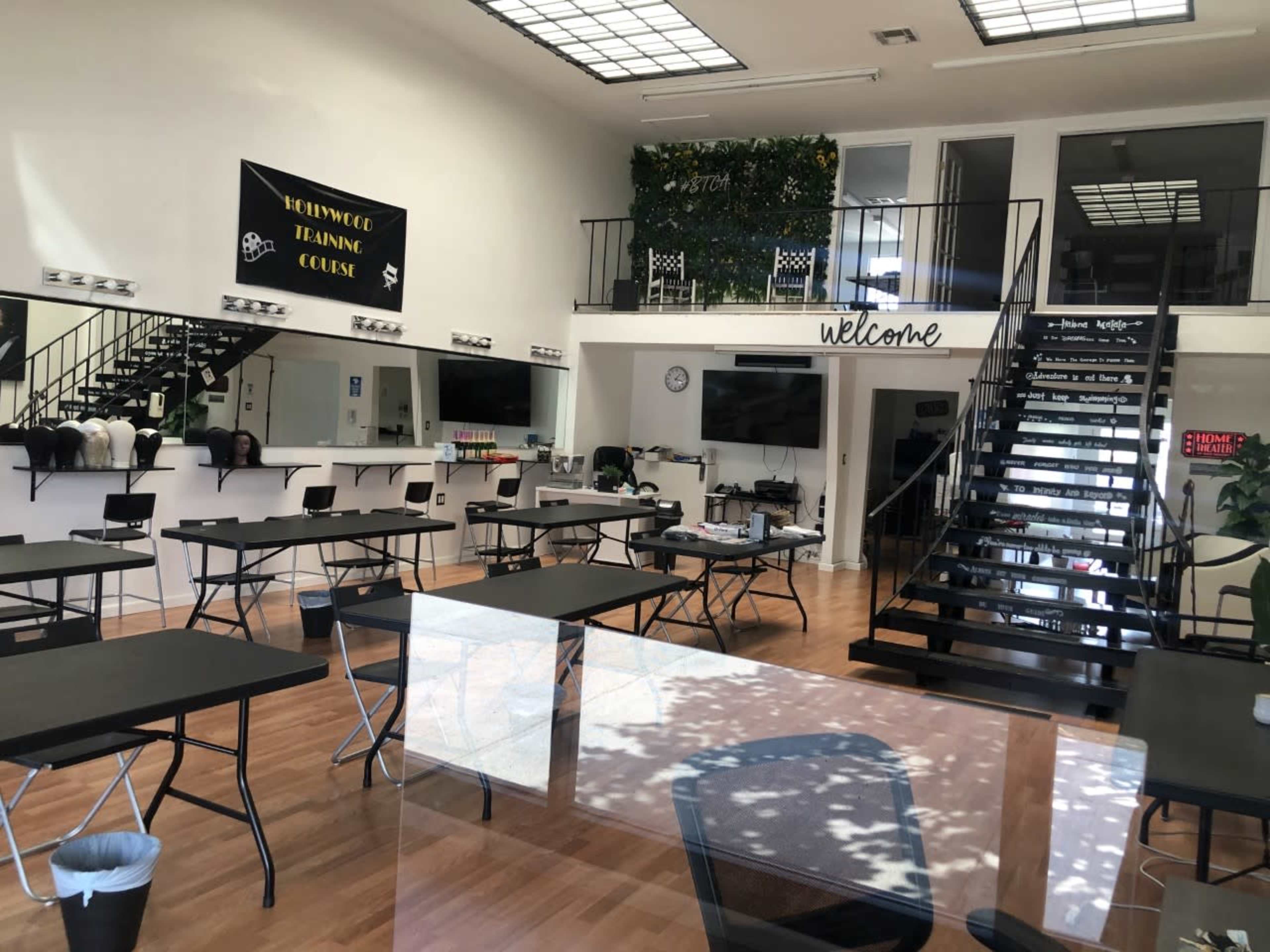 A spacious training classroom with black tables arranged throughout, a welcome sign on the wall, and a staircase leading to an upper level.