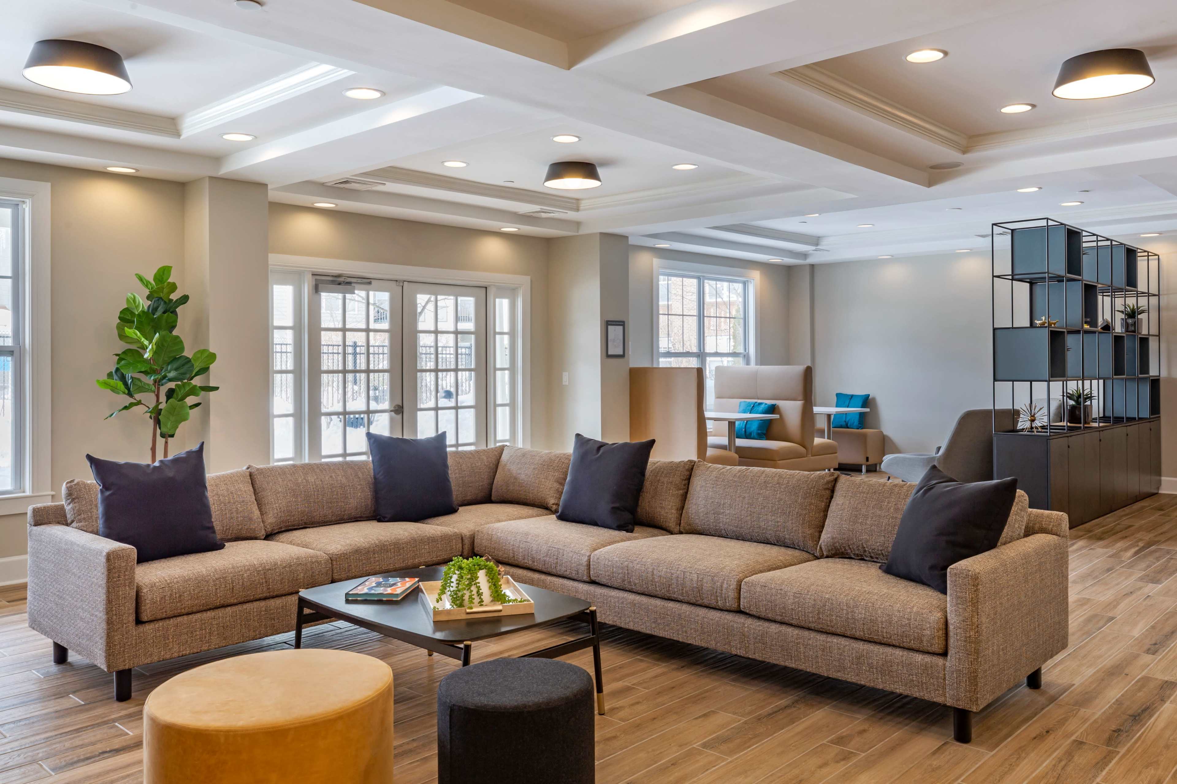 A modern seating area featuring a large sectional sofa with dark pillows, a coffee table, and a minimalistic bookshelf, all set in a well-lit room with large windows.