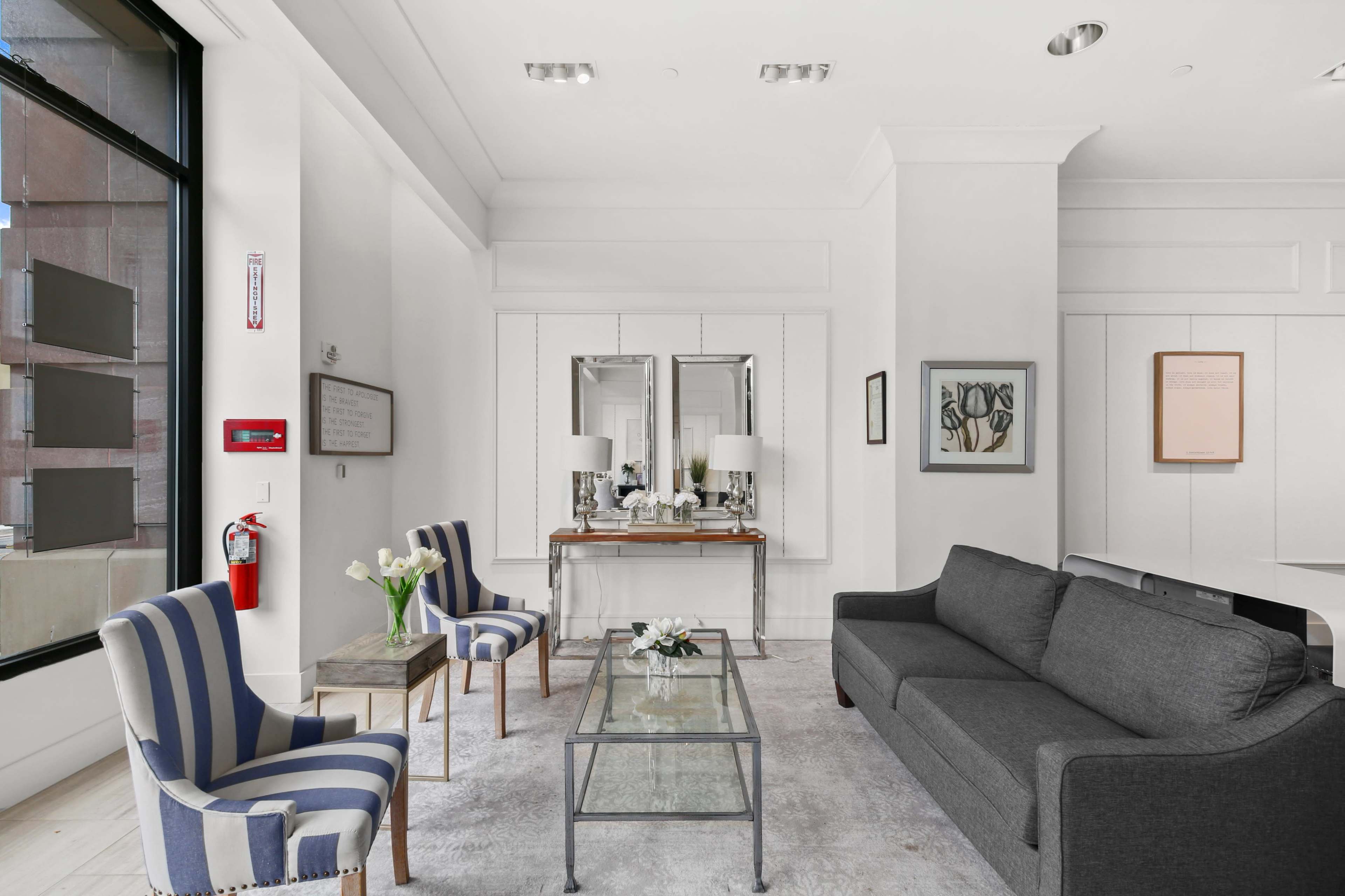 A modern living room with a gray sofa, two striped chairs, a glass coffee table, and a mirror reflecting the space.