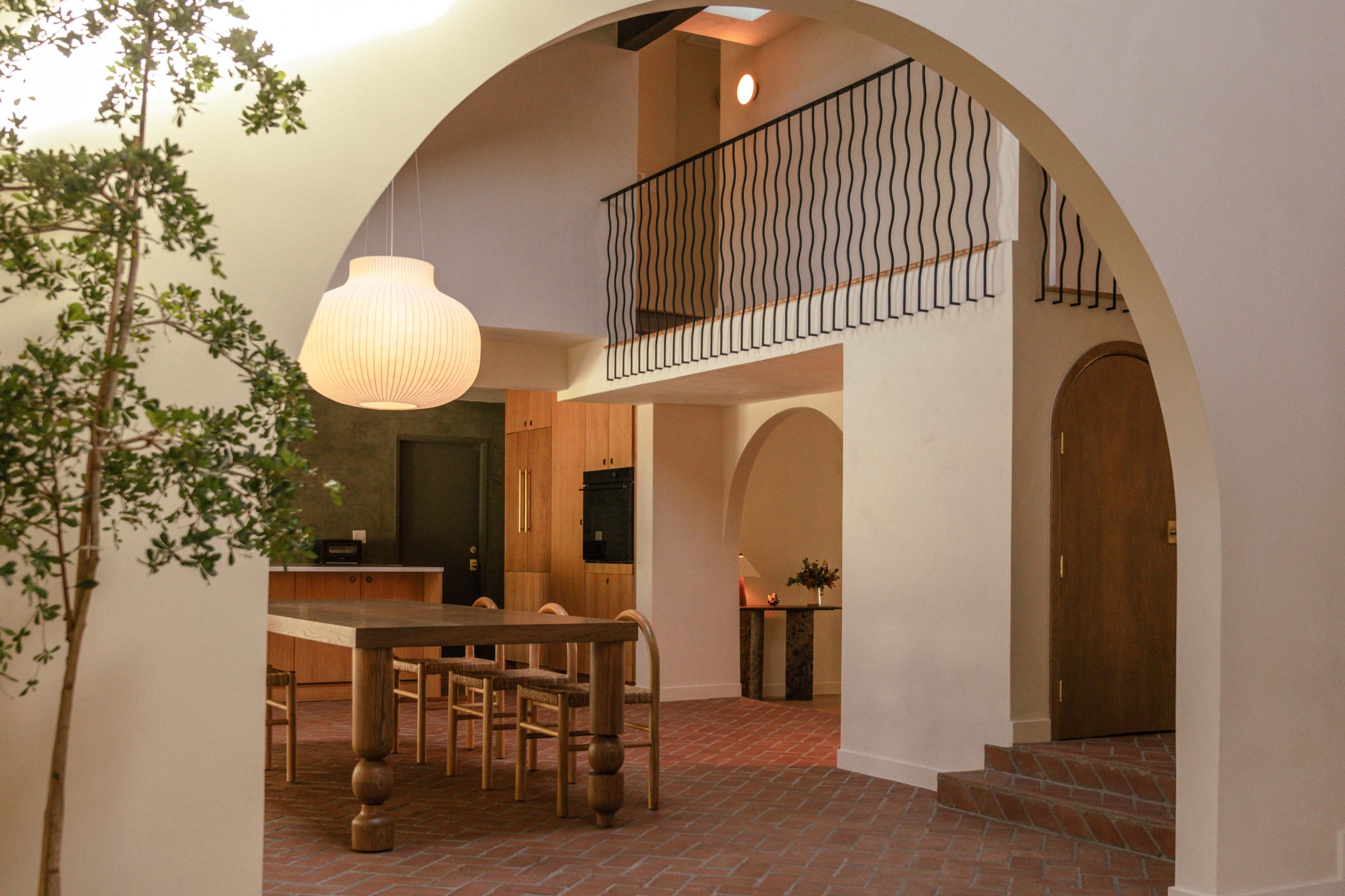 The image shows a modern interior space featuring arched openings, a wooden dining table, a spiral staircase, and a large pendant light.