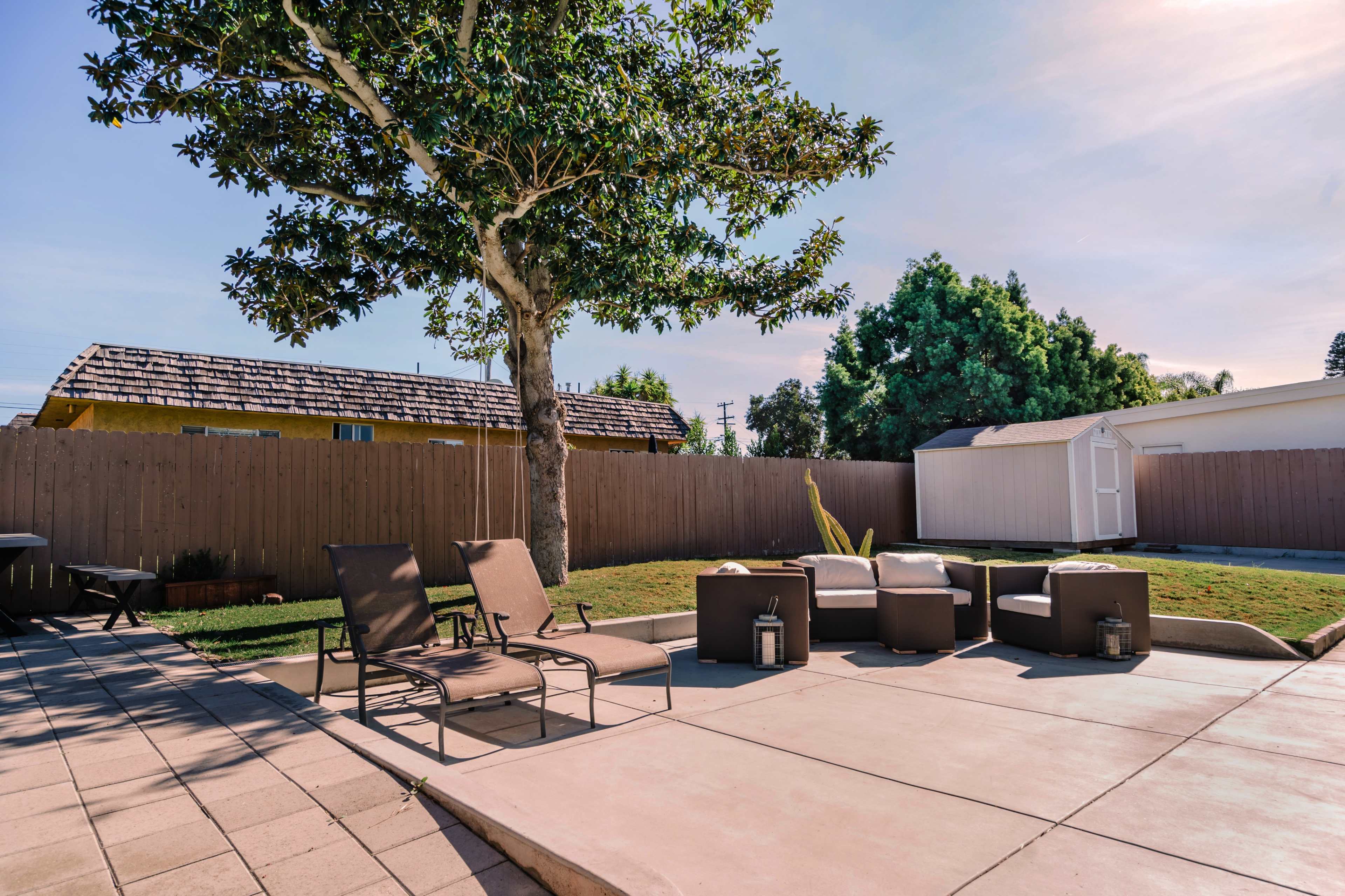 The image shows a backyard patio area with lounge chairs and a seating arrangement near a green lawn and a small shed in the background.