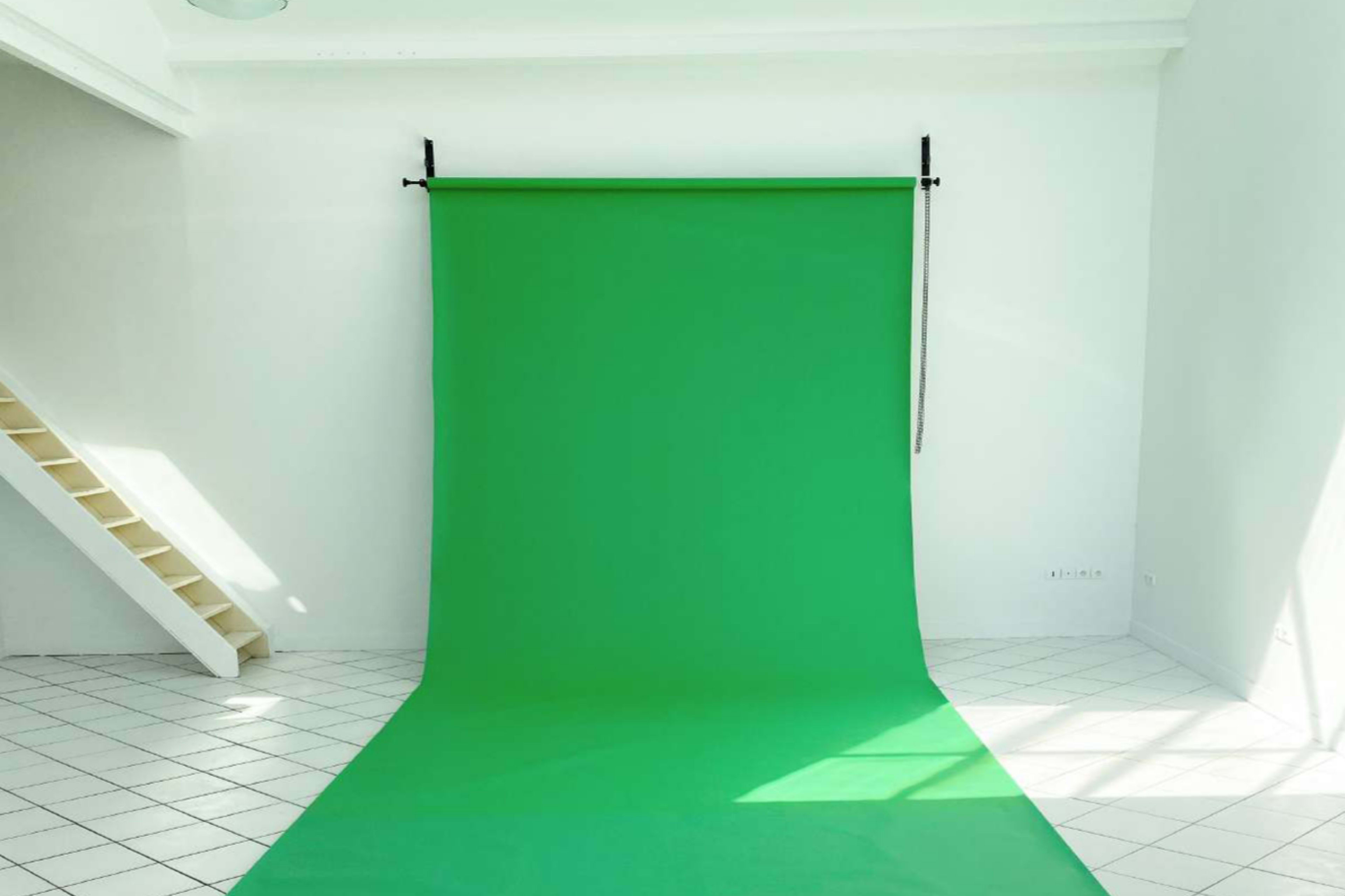 A green backdrop is set up in a brightly lit room with white walls and a staircase in the background.