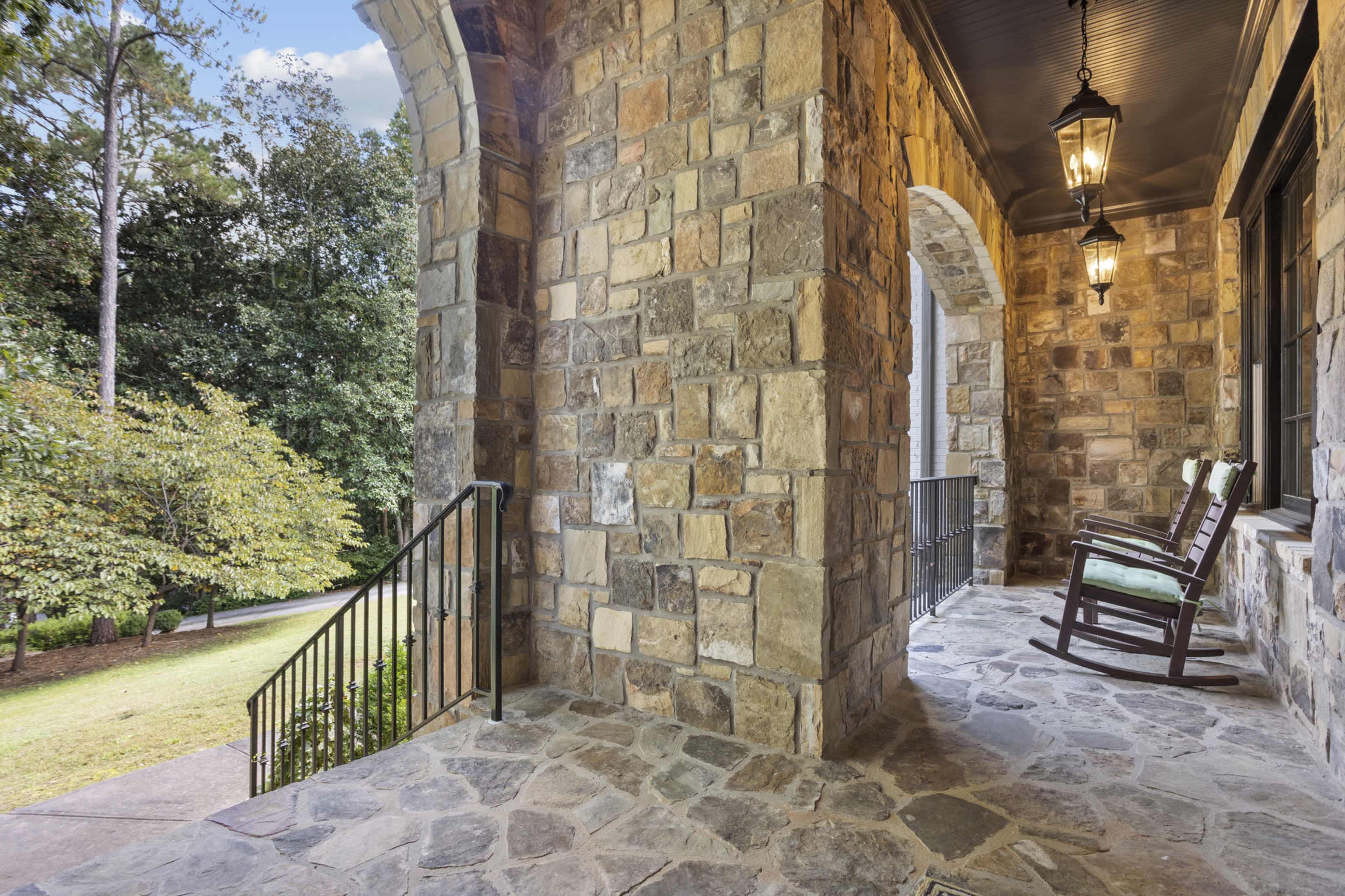A stone porch features two rocking chairs and overlooks a landscaped yard with trees.