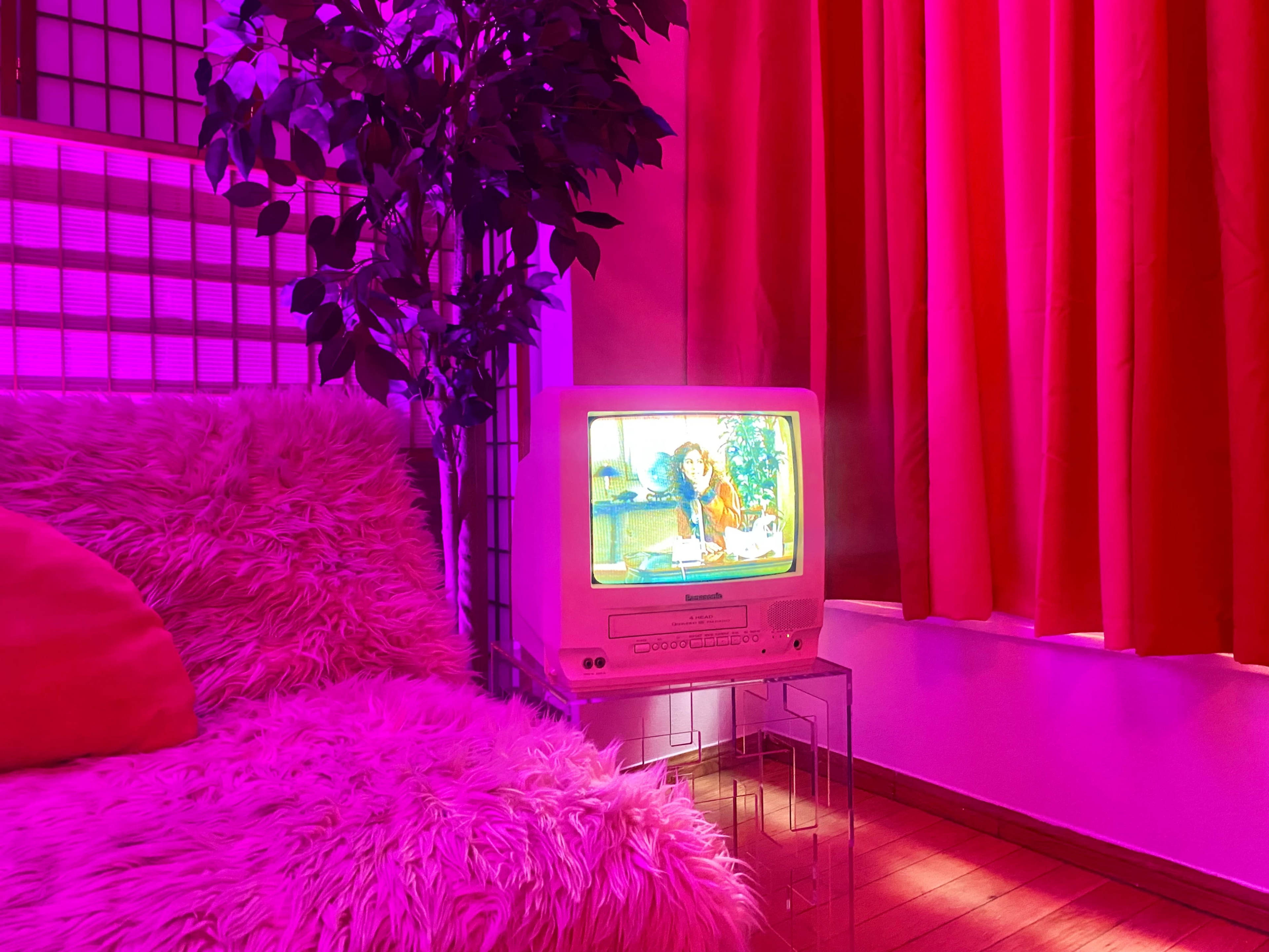 A small, retro television displaying a show sits on a clear stand next to a plush couch in a brightly lit room with pink curtains.