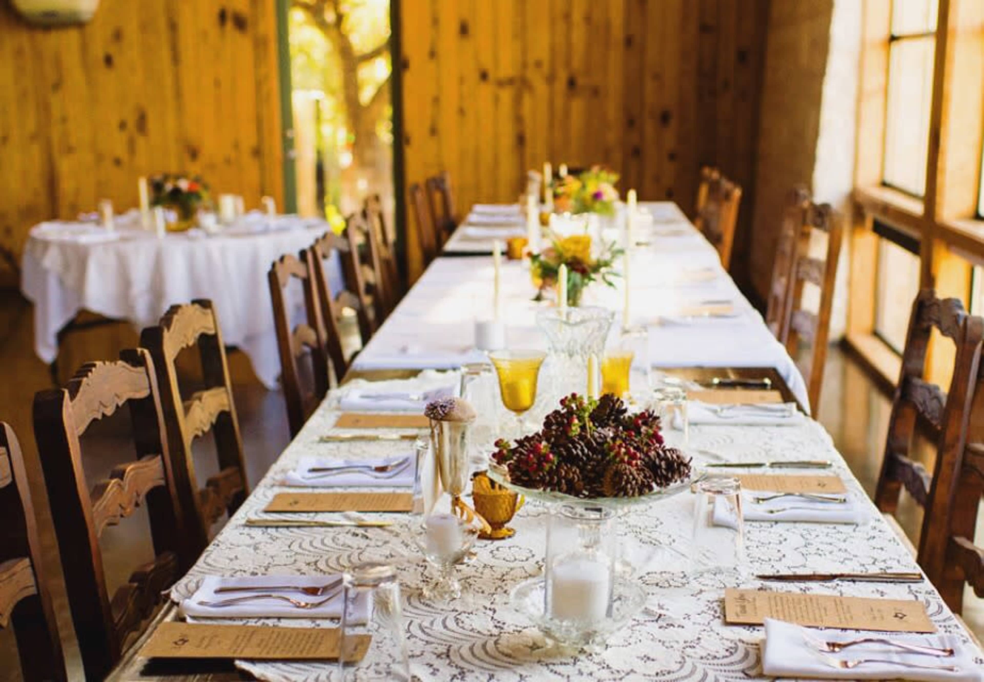 A long dining table is set with white tablecloths, ornate centerpieces, and neatly arranged place settings in a rustic wooden venue.