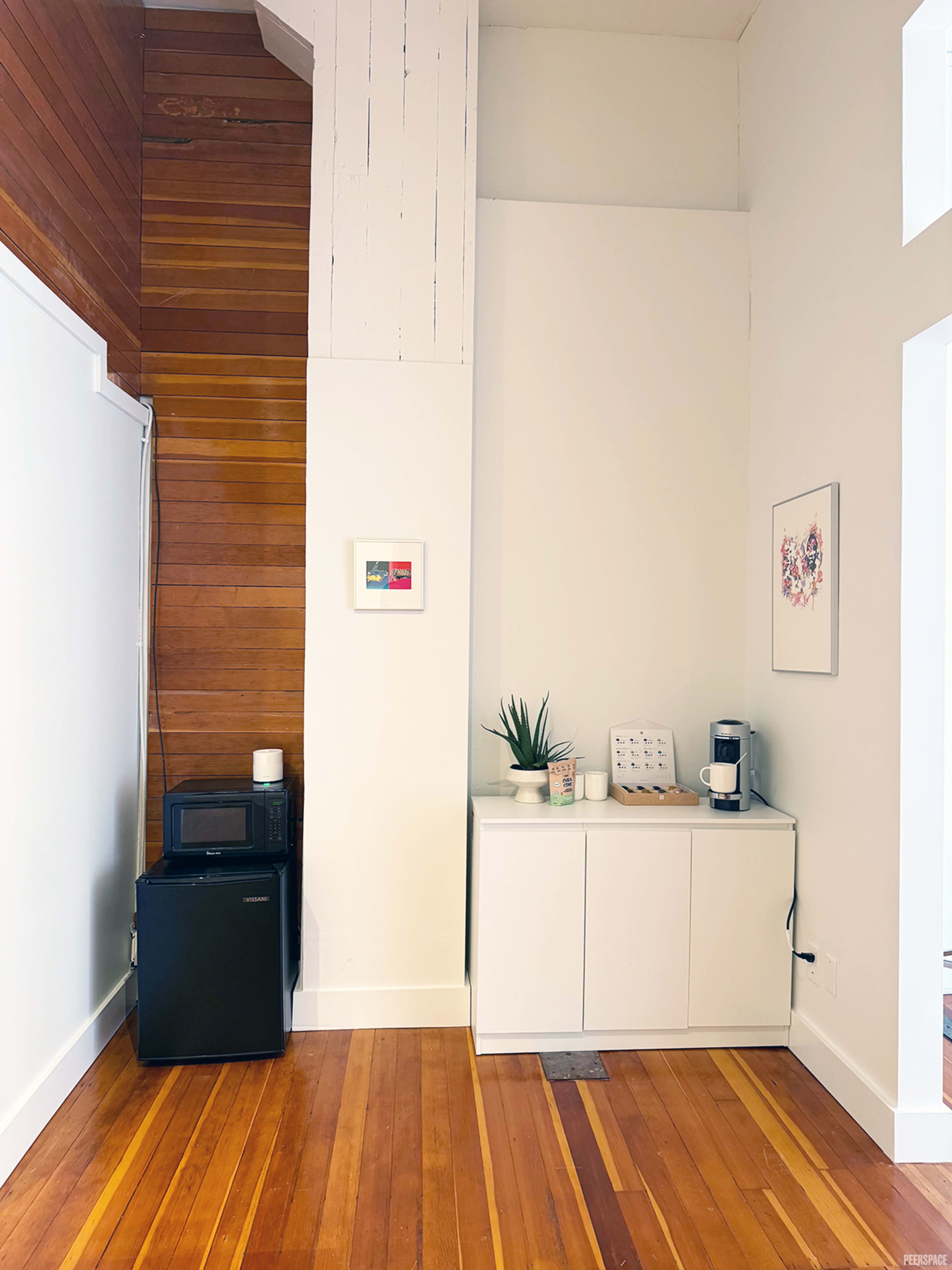 The image shows a modern, minimalist corner of a room featuring a black microwave and a white cabinet with a small plant and a coffee maker on top, against a backdrop of wooden walls and a light-colored ceiling.
