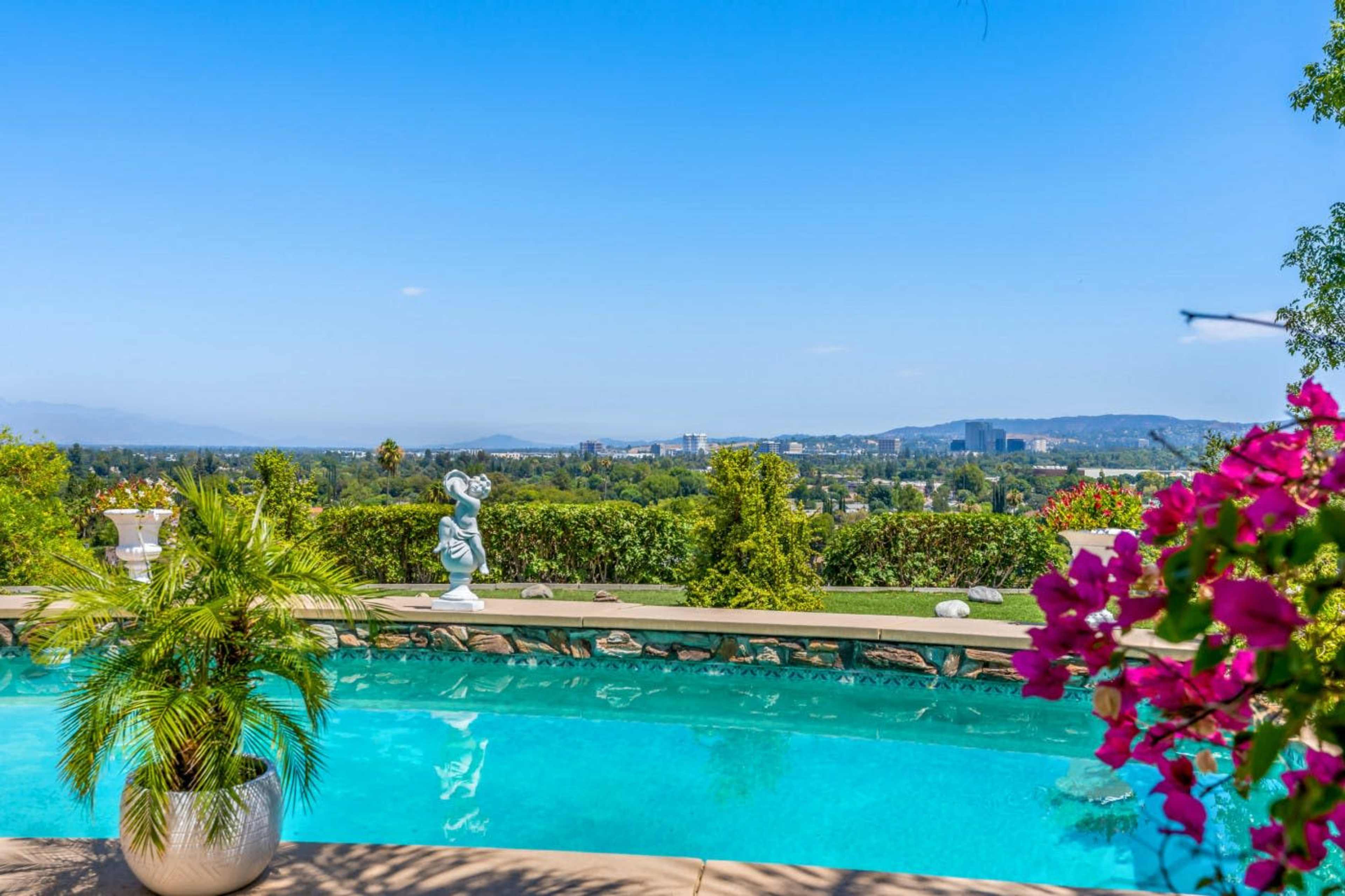 The image shows a swimming pool surrounded by greenery and a view of distant buildings and mountains under a clear blue sky.