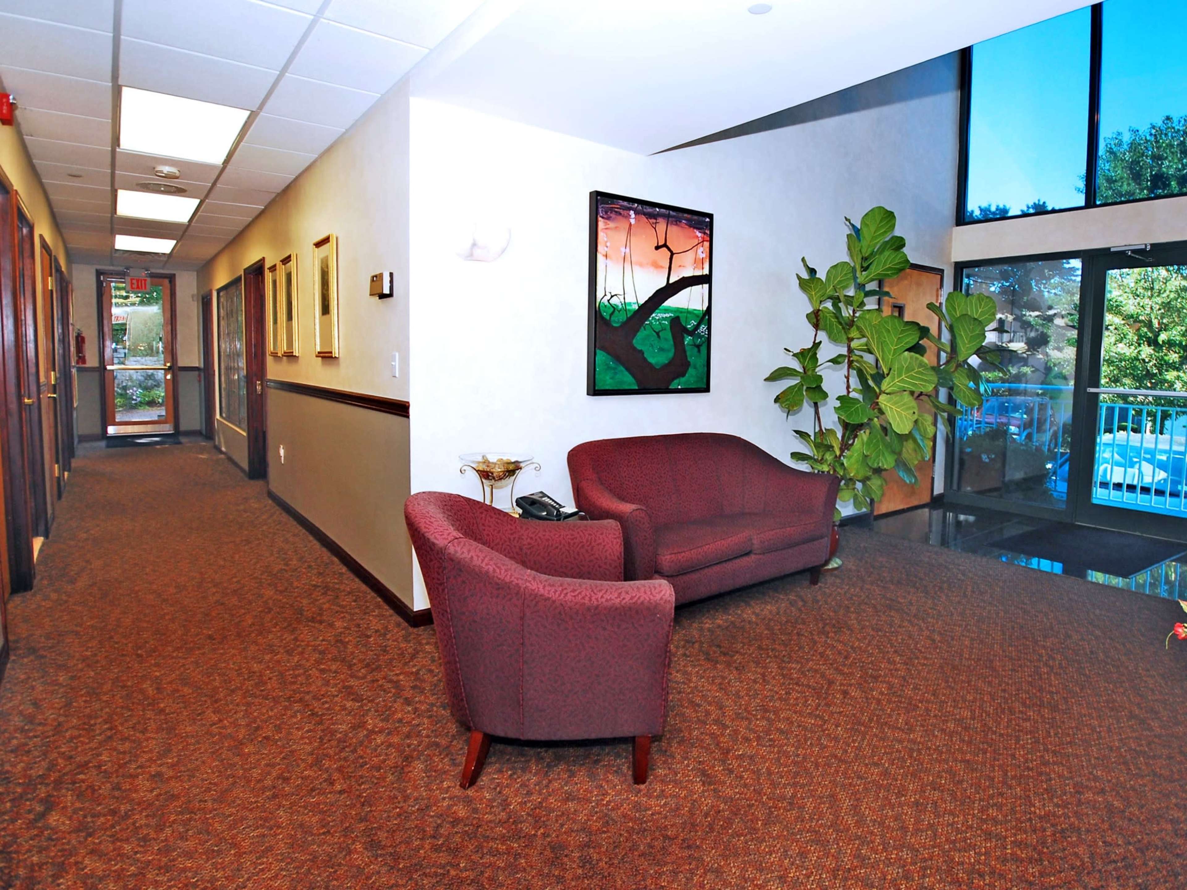 The image shows a hallway with carpeted flooring, a pair of maroon couches, and a large window that provides natural light and a view of outdoor greenery.