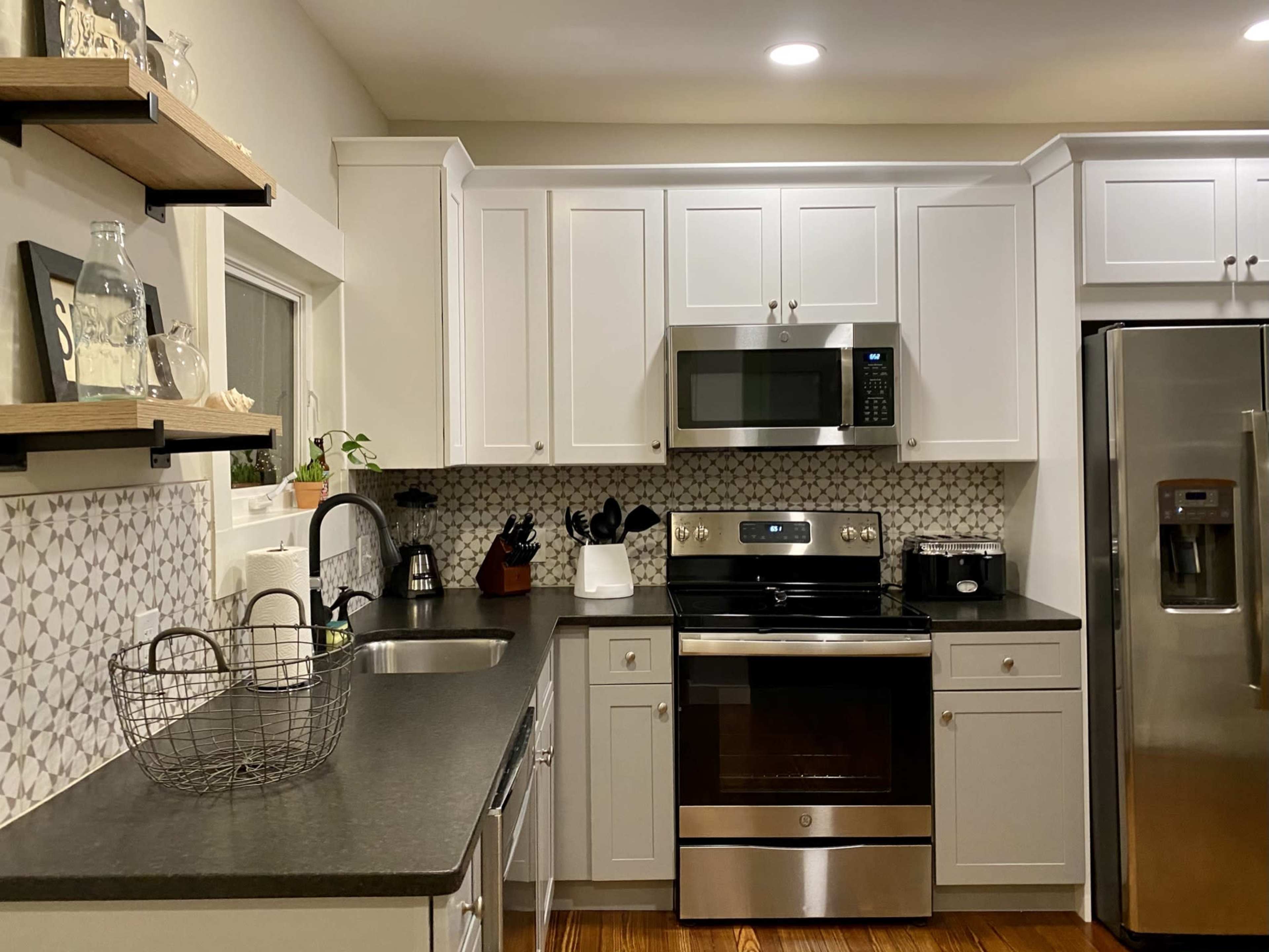 The image shows a modern kitchen featuring white cabinetry, a stainless steel microwave and oven, a black countertop, and patterned backsplash tiles.