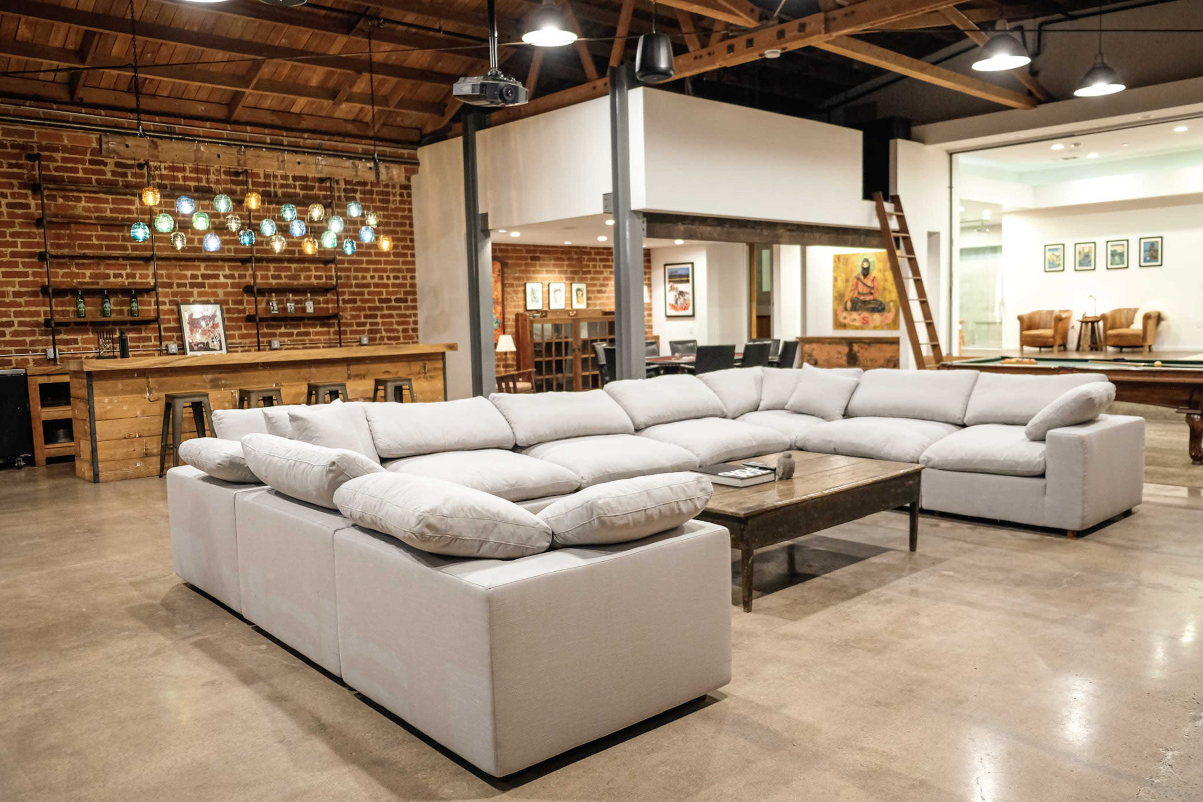 The image shows a spacious interior with a large, modular white sectional sofa and a wooden coffee table, surrounded by exposed brick walls and modern lighting fixtures.