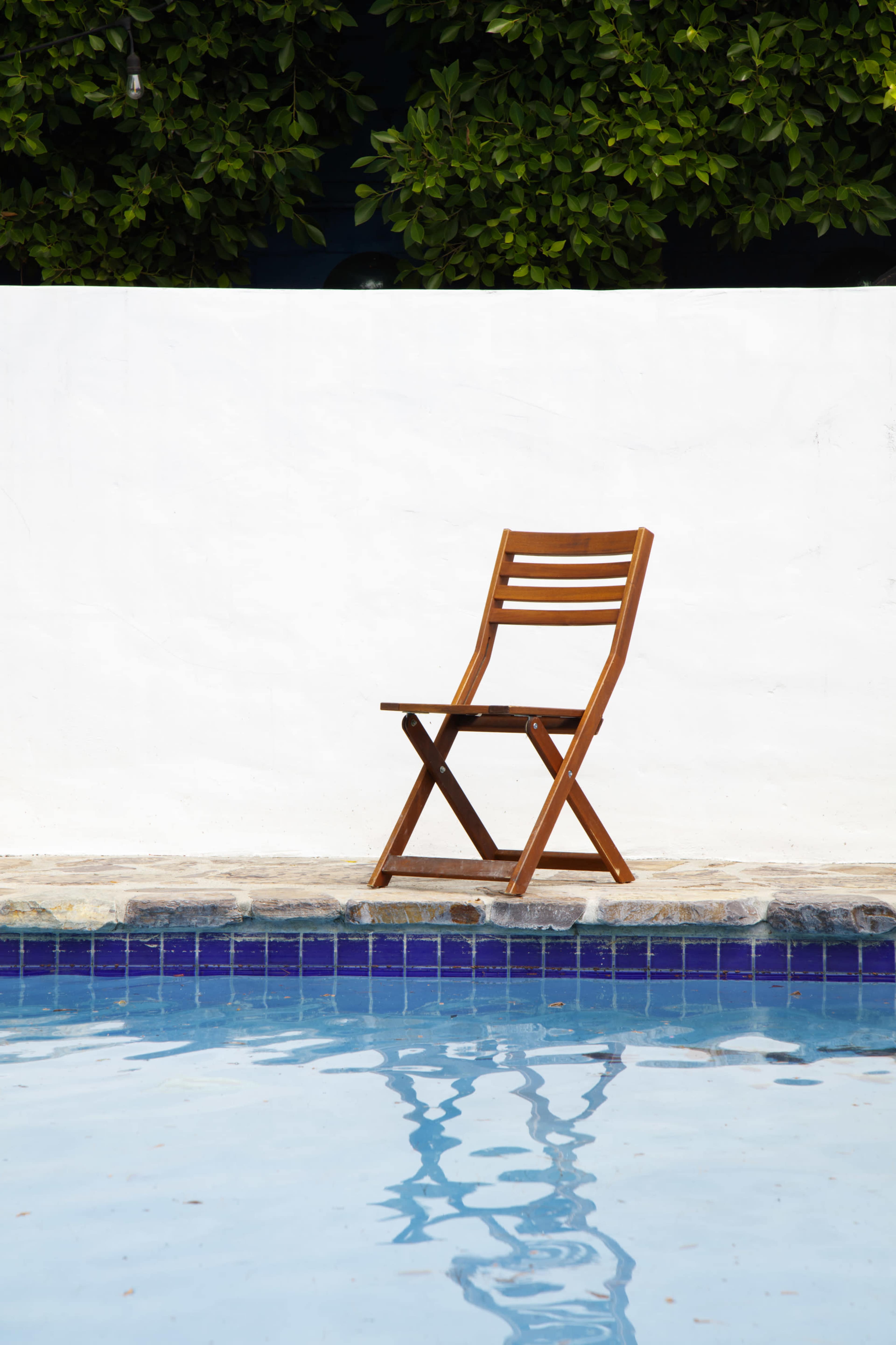 A wooden folding chair sits beside a swimming pool, reflecting in the water below.