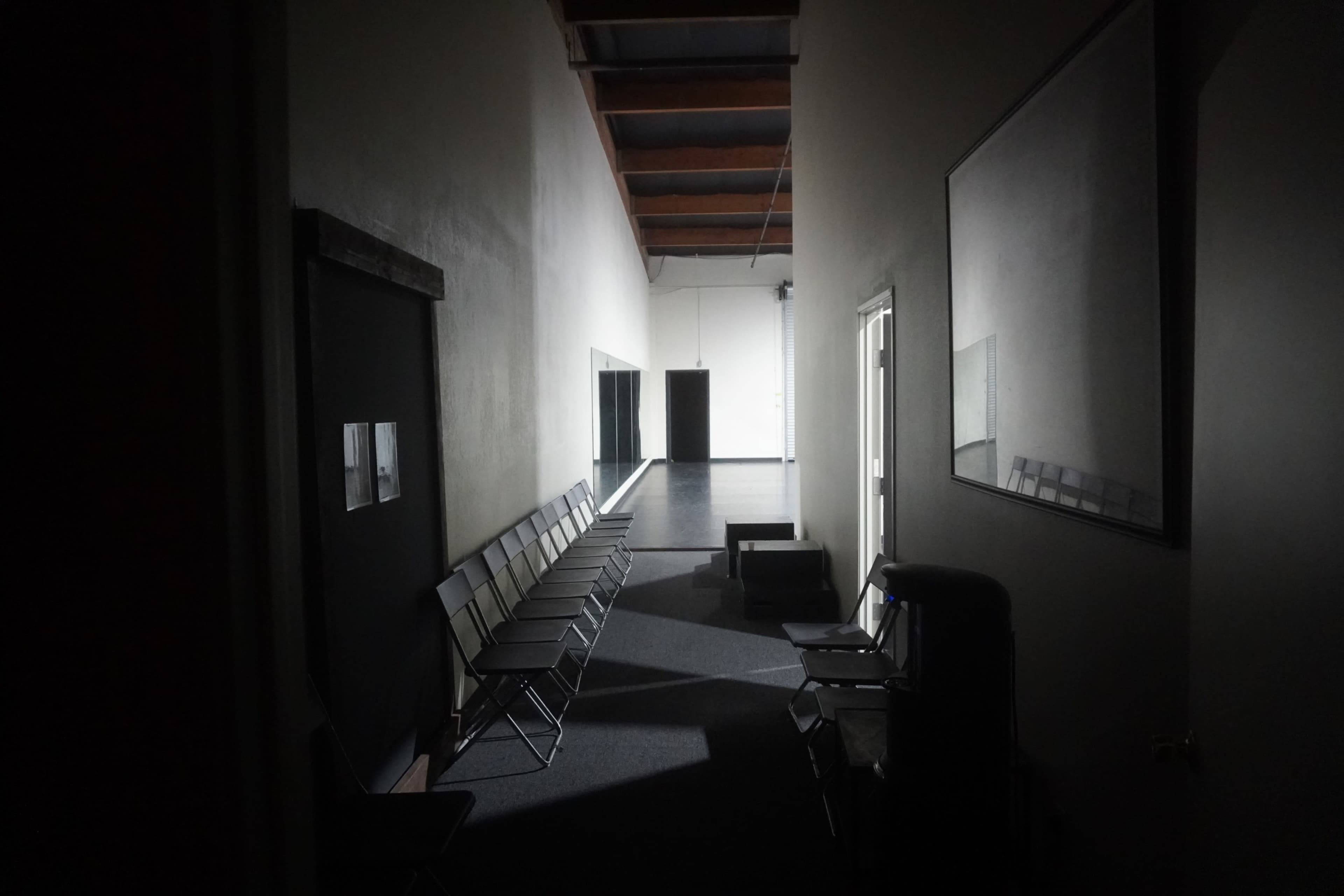 A dimly lit corridor features a row of metal chairs along one side and doors leading to adjoining rooms.
