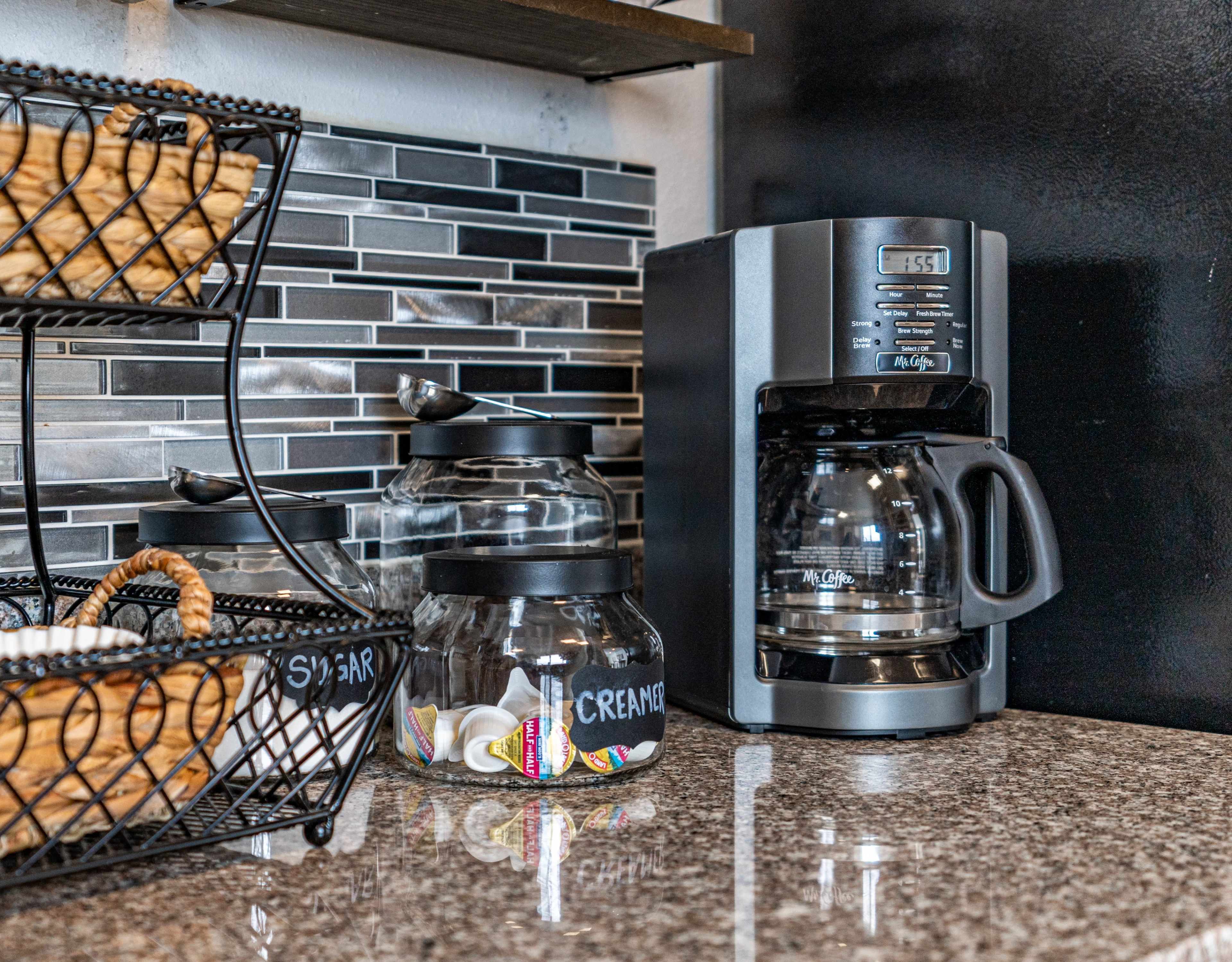 A coffee maker is positioned next to a granite countertop with jars labeled for sugar and cream, and a wire basket containing pastries is above.