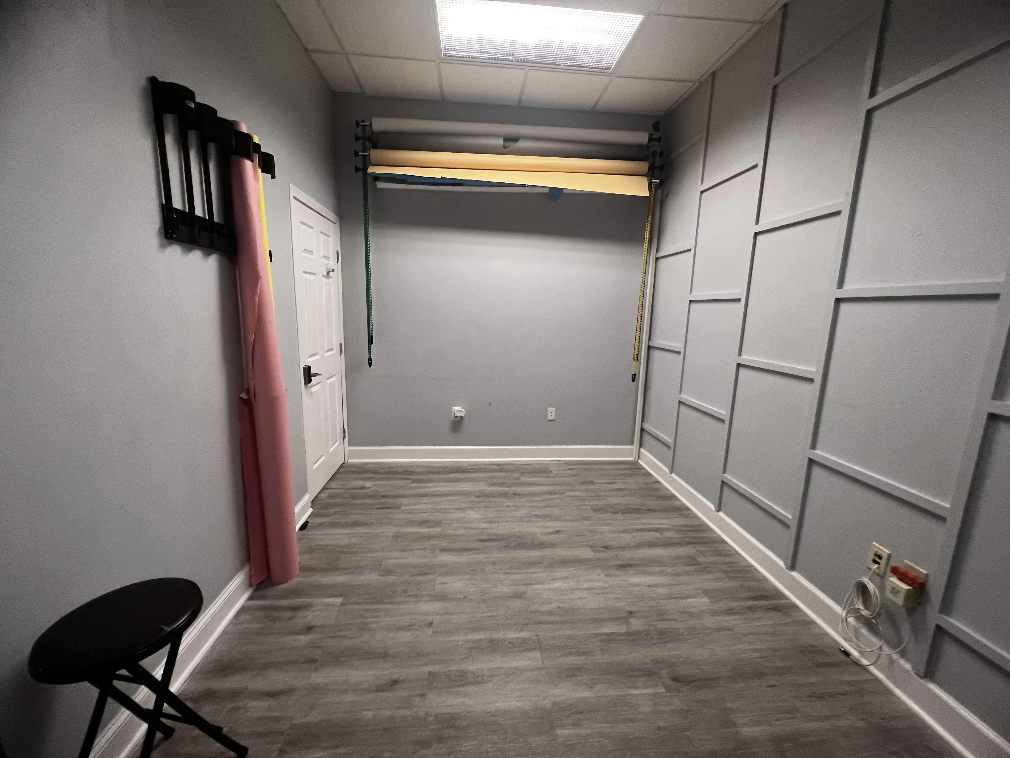 A sparse room with gray walls, a door, a small black stool, and rolls of colored backdrop paper mounted on a wall.