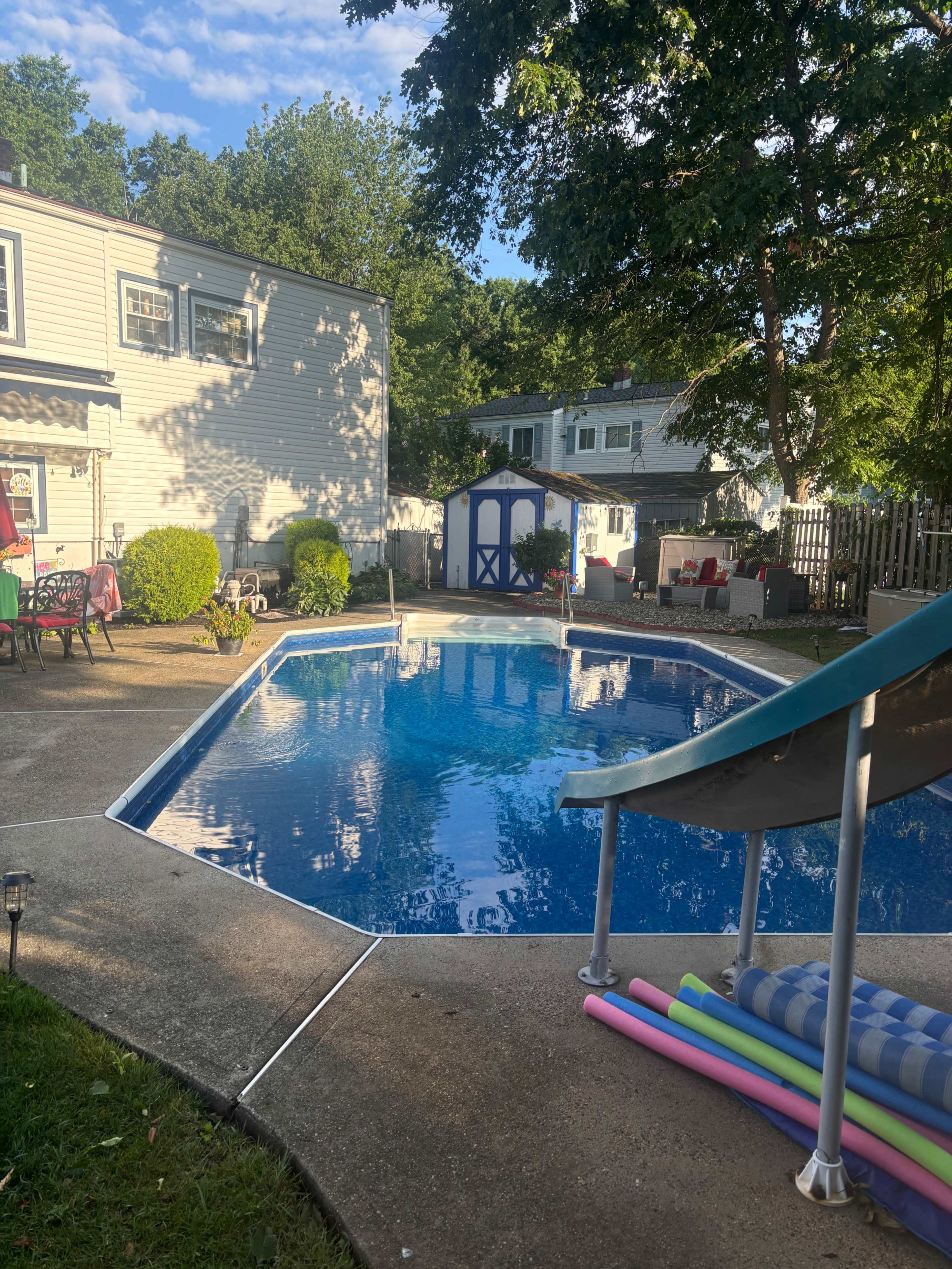A backyard swimming pool with a blue water slide, surrounded by grass, patio furniture, and colorful pool noodles.