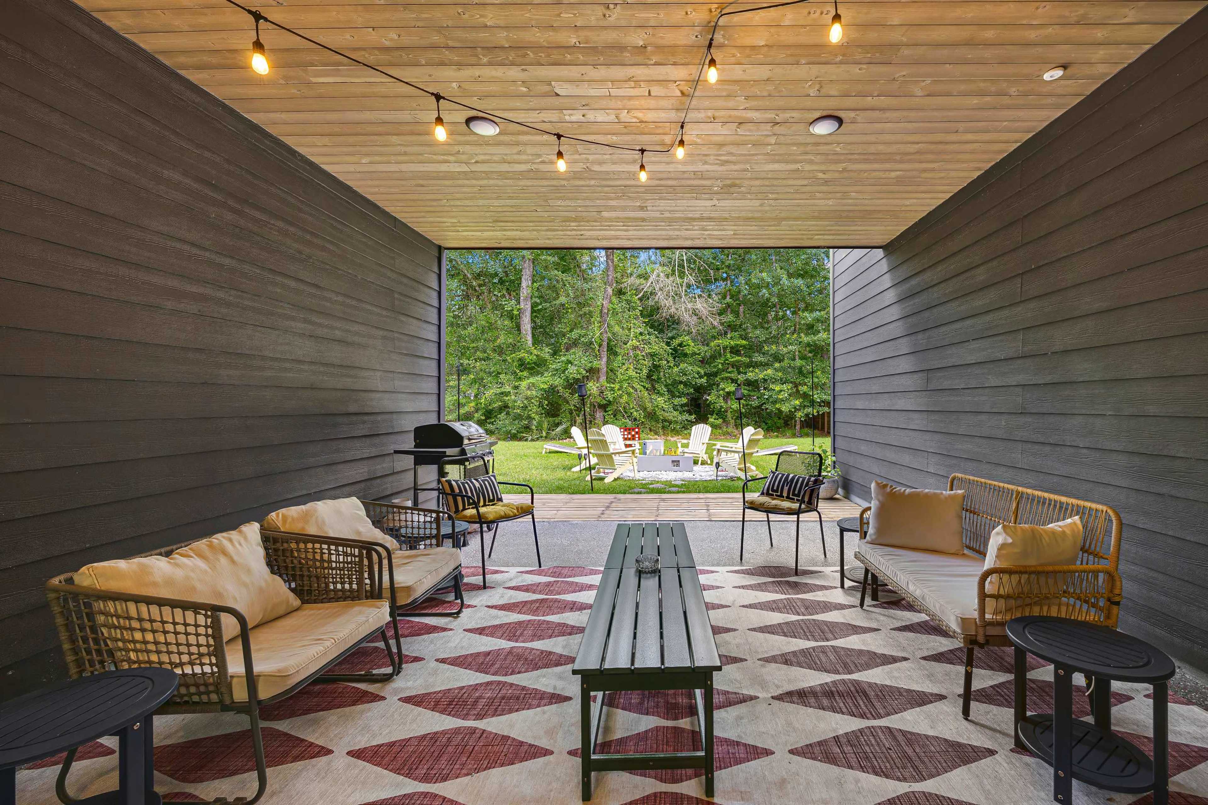 The image shows a covered outdoor seating area with string lights, surrounded by greenery and a patio with lounge chairs in the background.