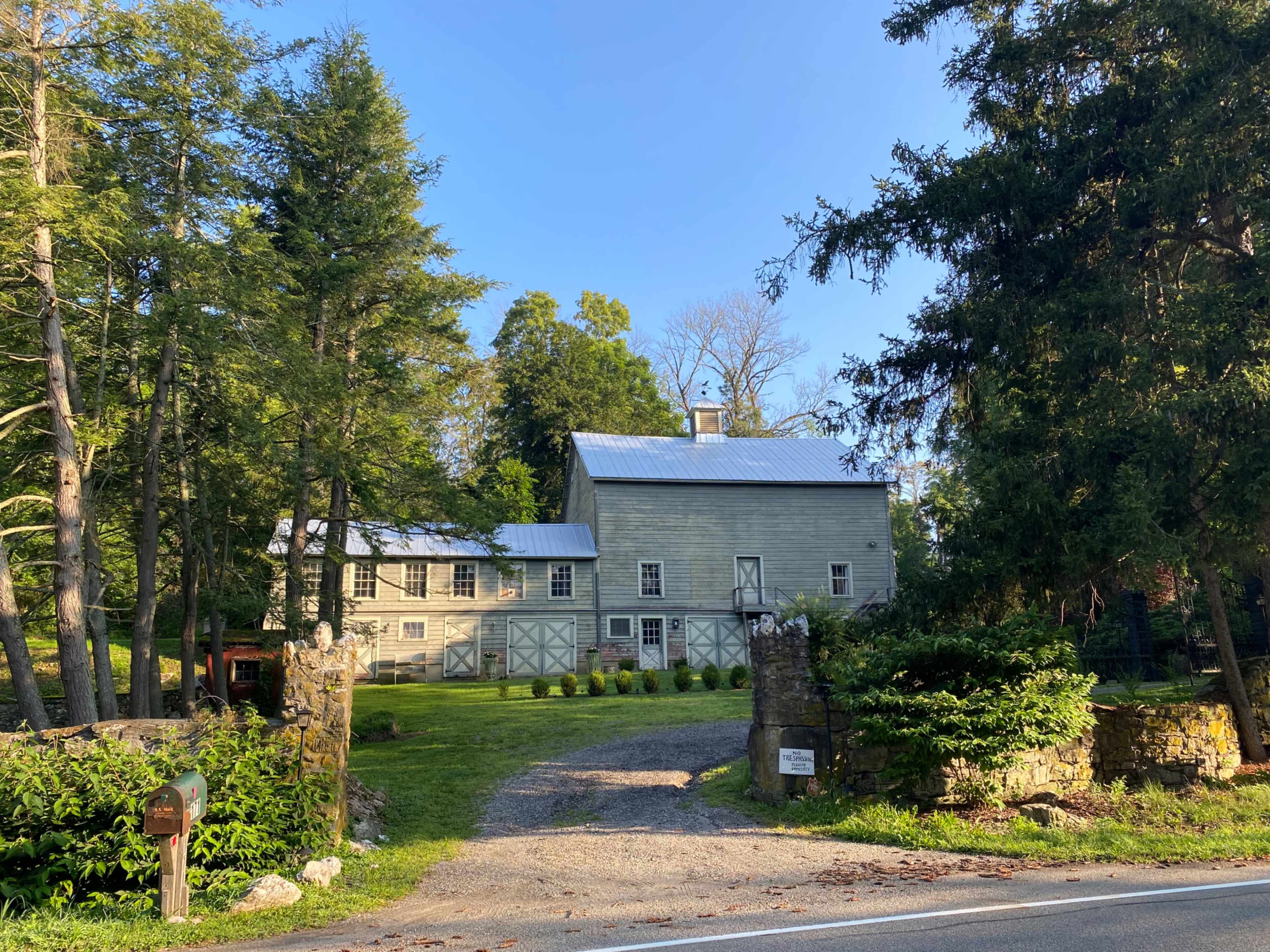 A gray wooden barn-style house is set back from a gravel driveway surrounded by trees and greenery.