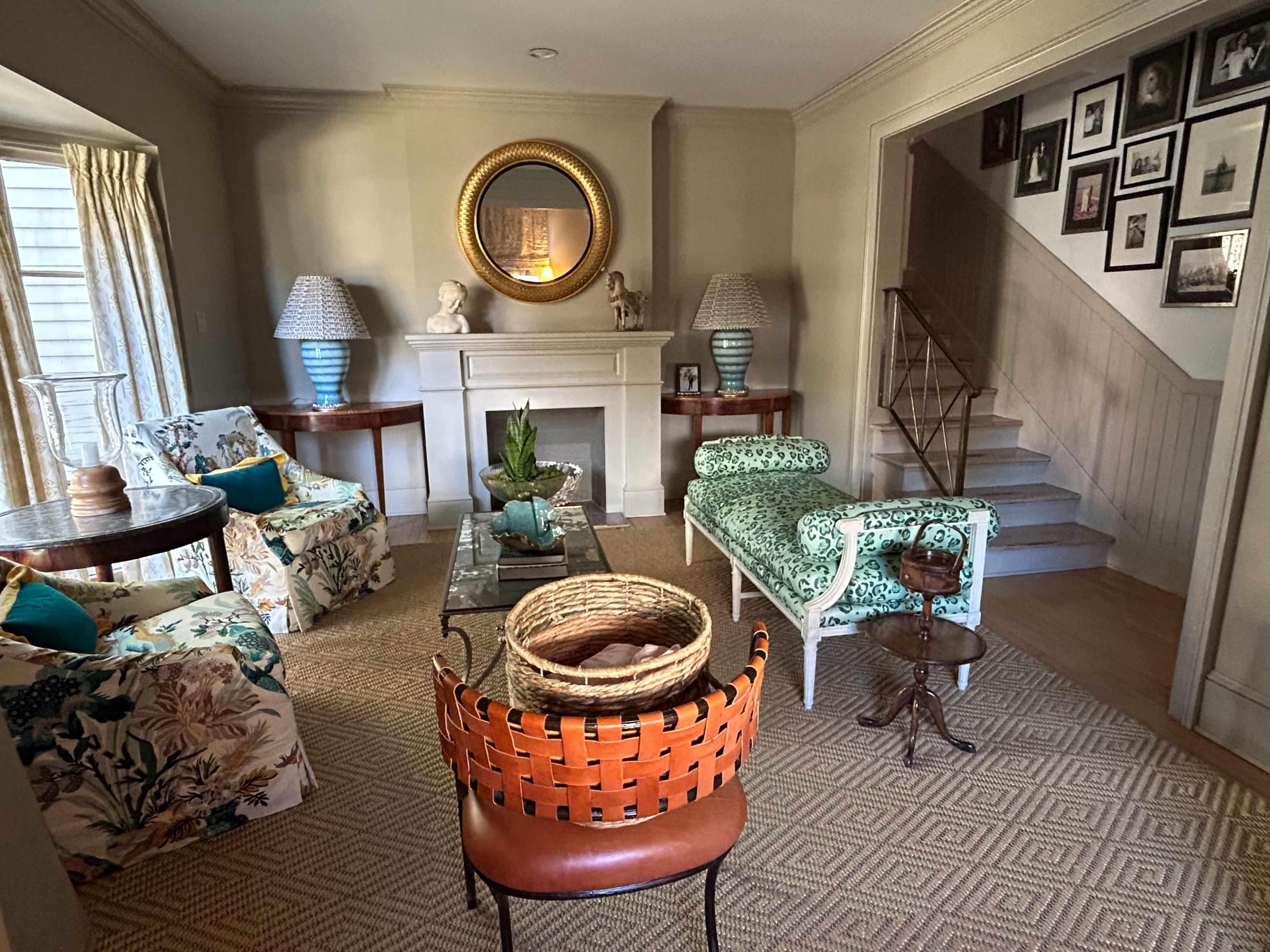 The image shows a living room with patterned chairs, a fireplace, a large mirror, and a staircase leading to an upper level.