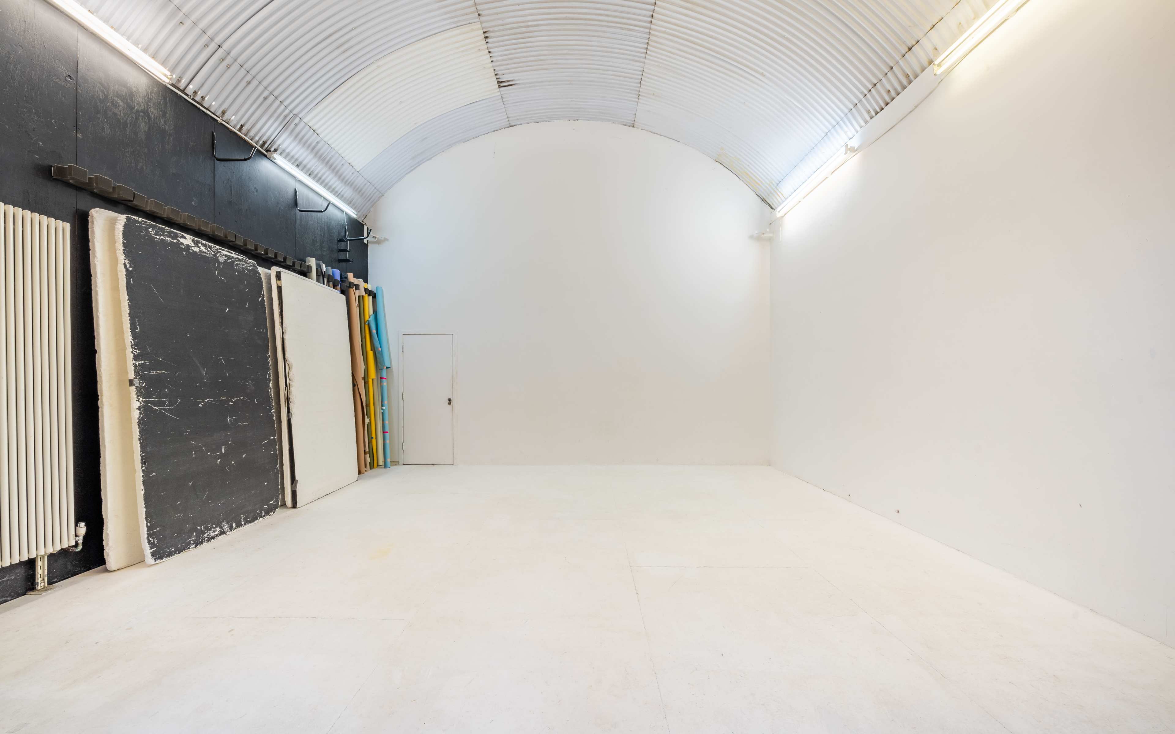 An empty room with a curved ceiling, featuring white walls, a door, and stacked materials against one side.