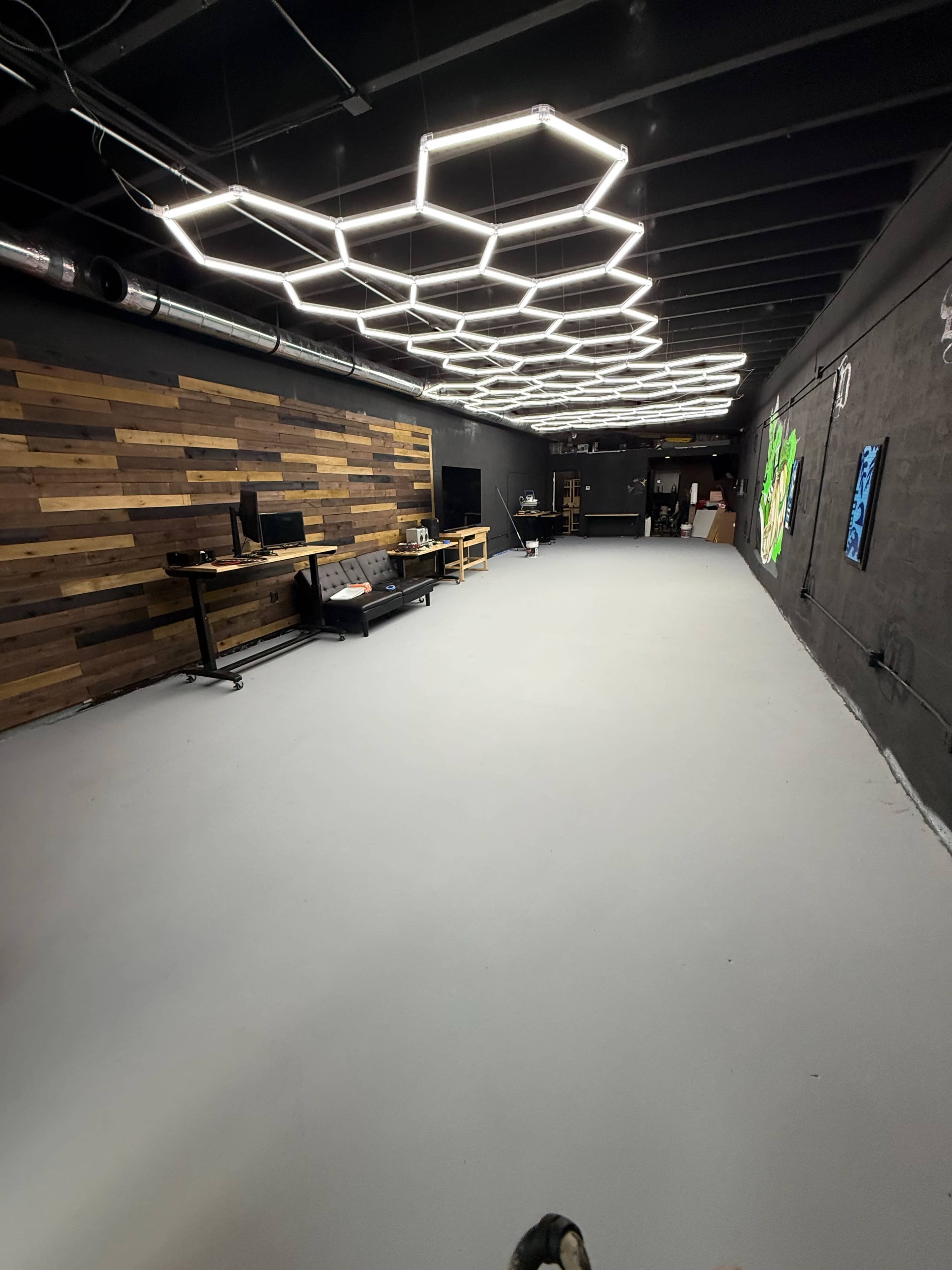 The image shows a spacious, modern interior with a hexagonal light fixture, wooden accent wall, and minimal furnishings including desks and artwork on the walls.