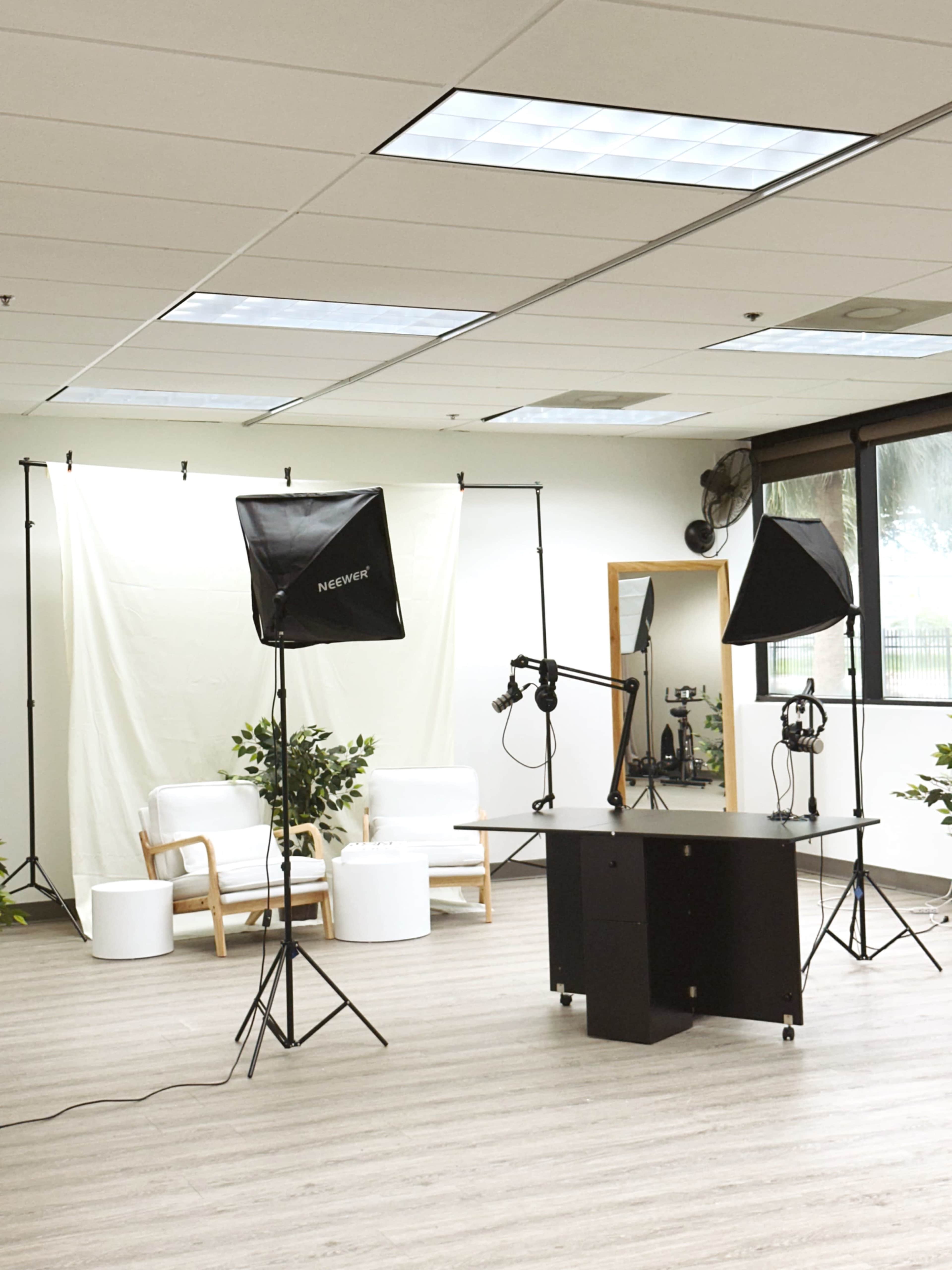 The image shows a professional studio setup with a white backdrop, lighting equipment, and a black table, surrounded by potted plants and a seating area.