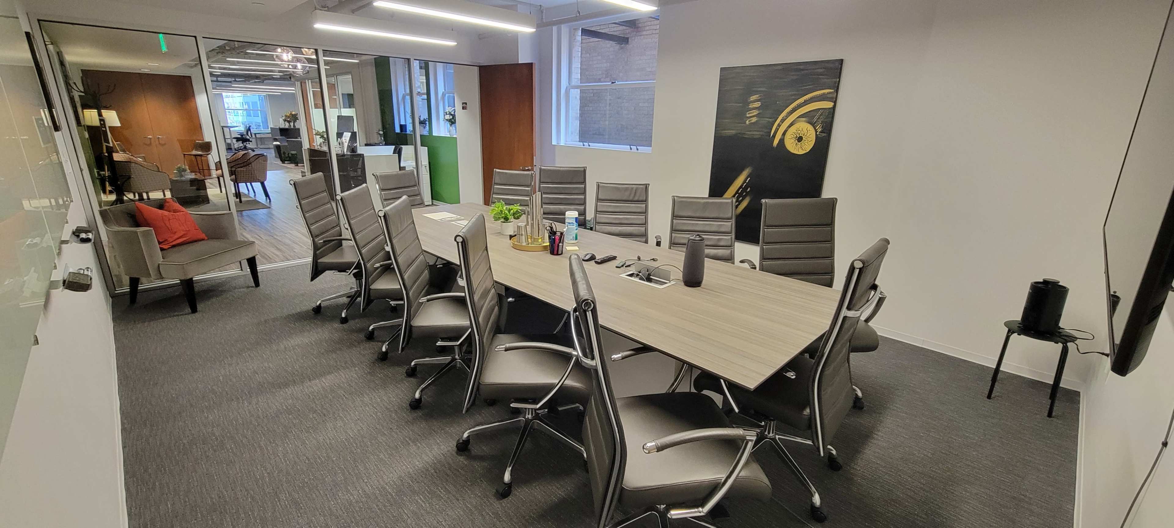 A modern conference room is set up with a long wooden table surrounded by ergonomic chairs, equipped with a whiteboard and a television on the wall.