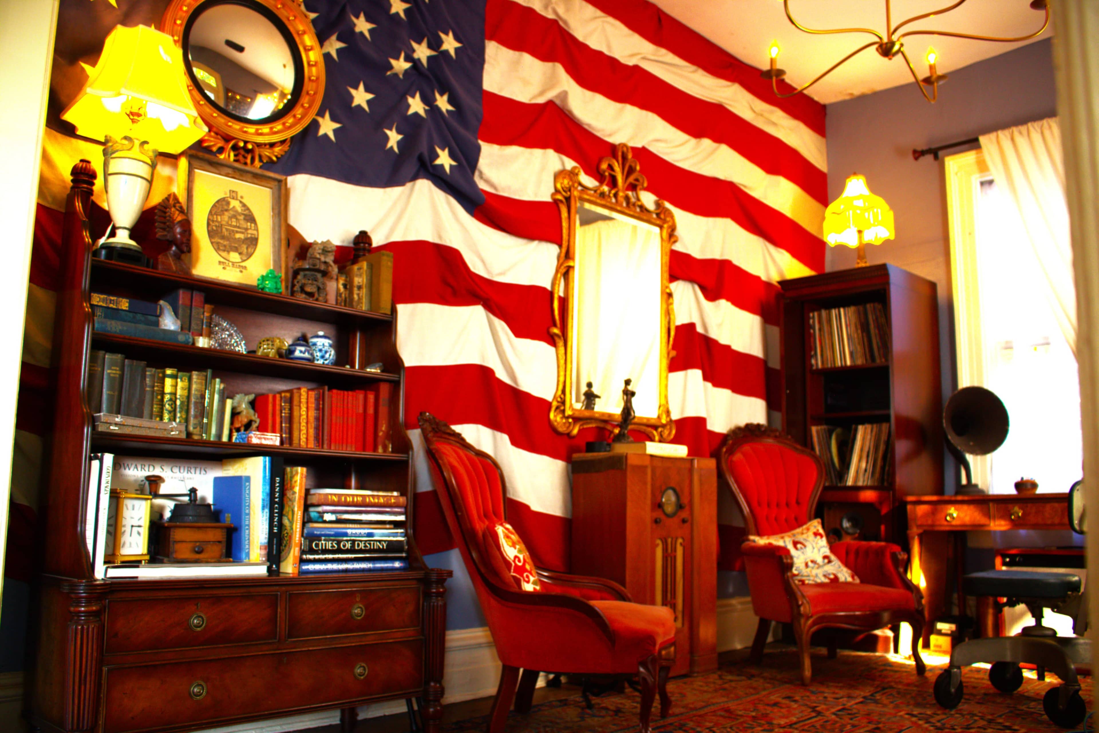 The image shows a room decorated with an American flag wall, featuring vintage furniture including a mirror, bookshelves, and two red chairs.