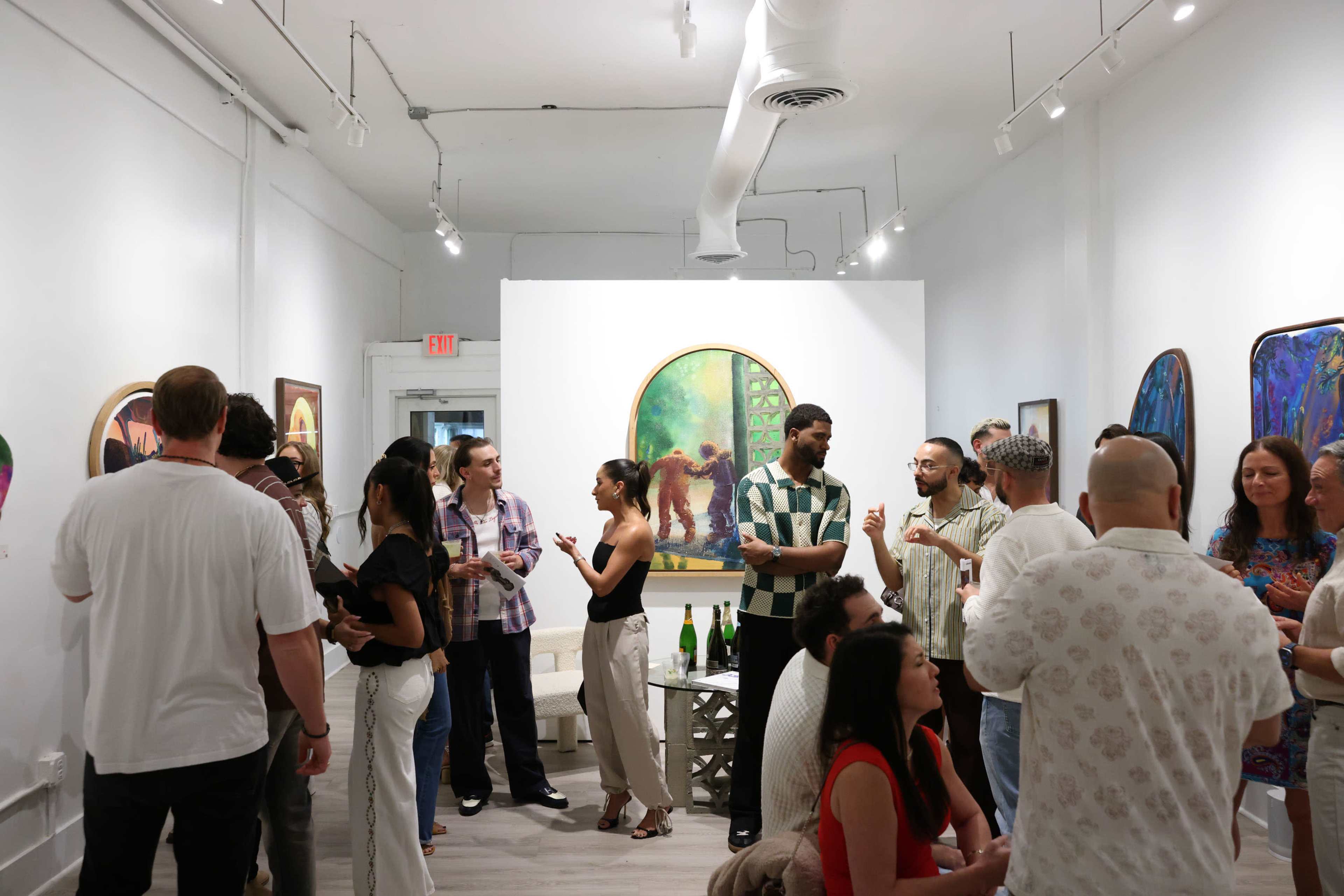 A group of people socialize in an art gallery, surrounded by colorful paintings displayed on white walls.