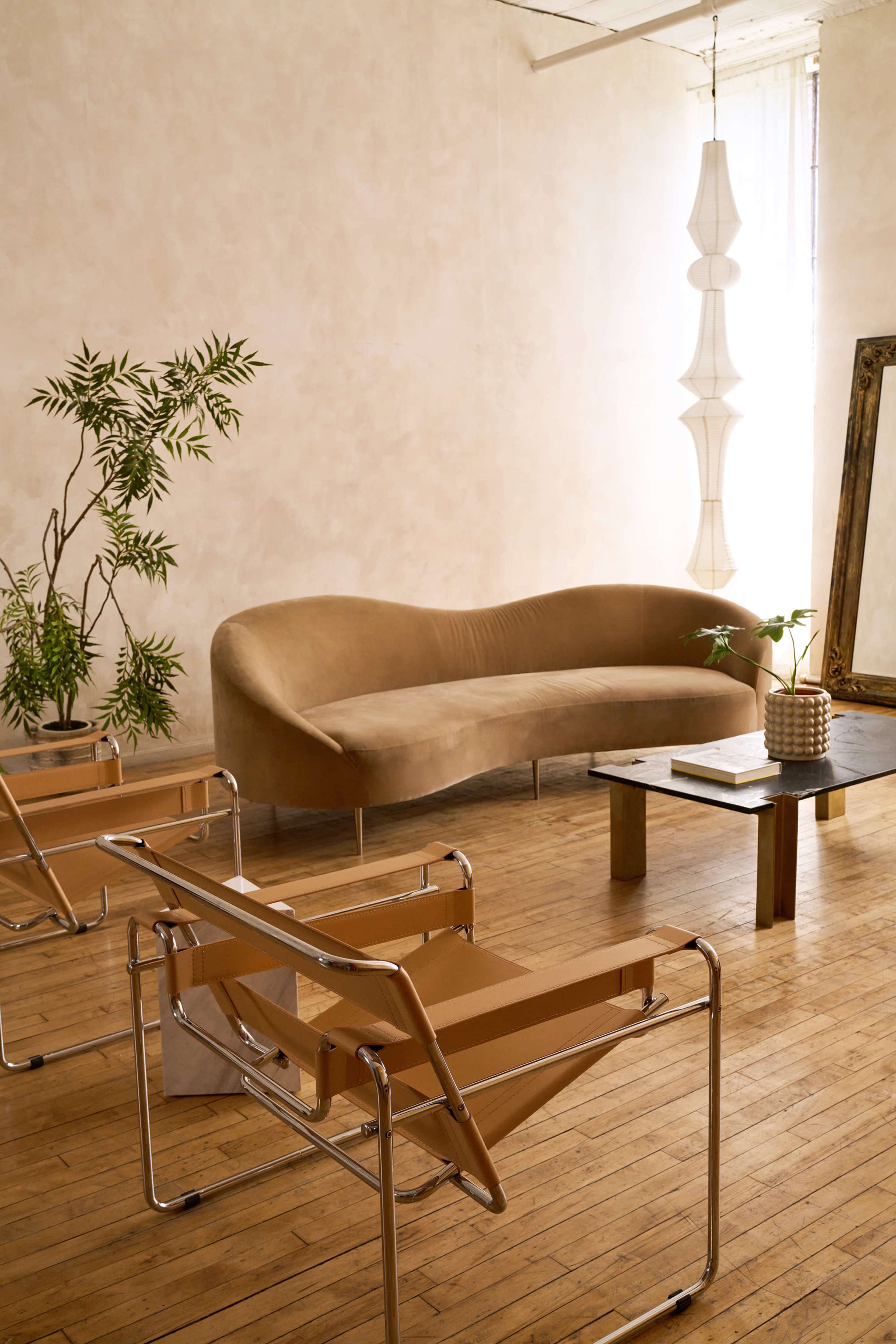 The image depicts a modern living space featuring a curved beige sofa, two minimalist chairs, a wooden coffee table, and a potted plant, all set against a light-colored wall.