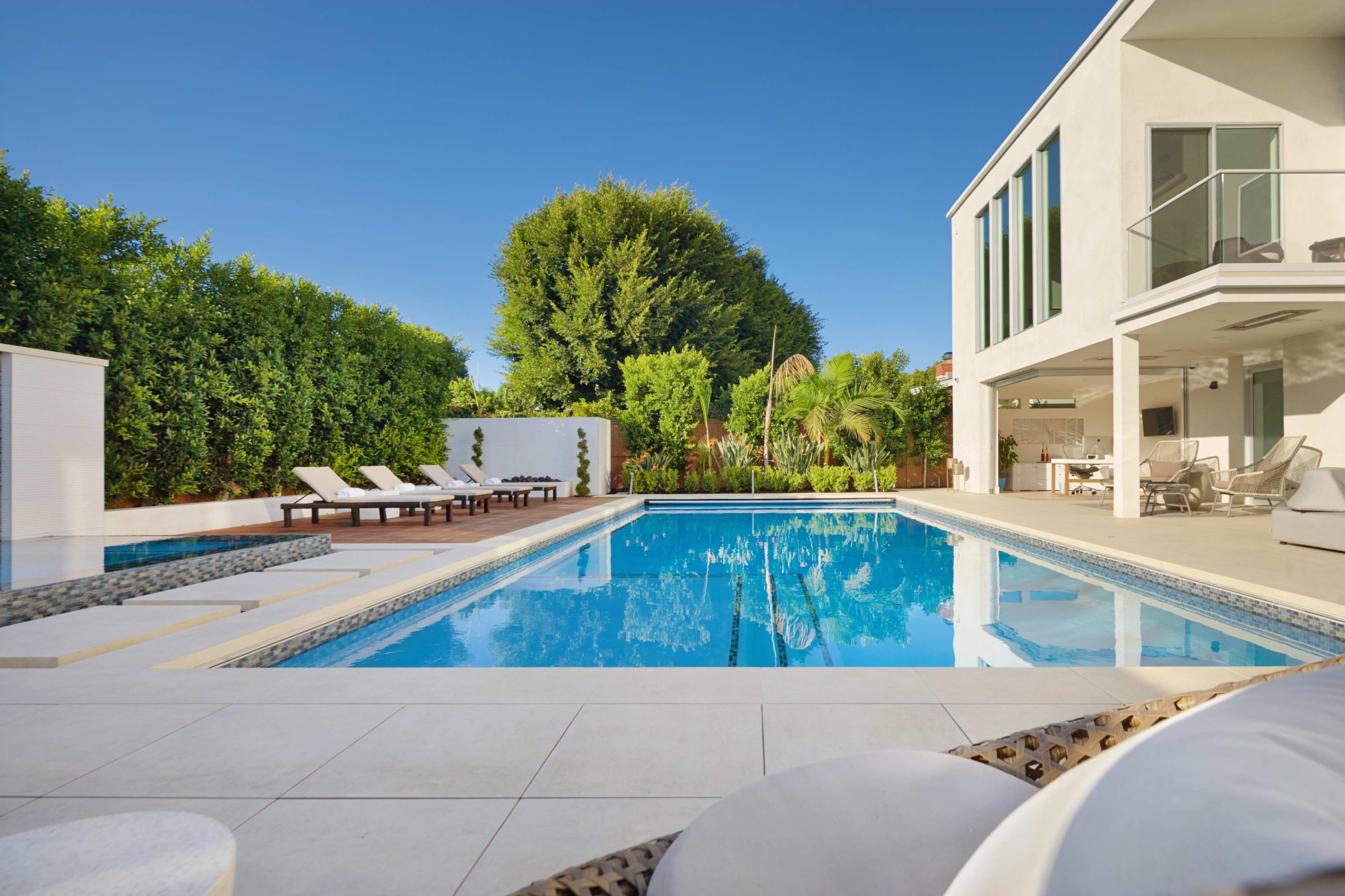 A modern backyard pool area features a large rectangular pool surrounded by lounge chairs and lush greenery.