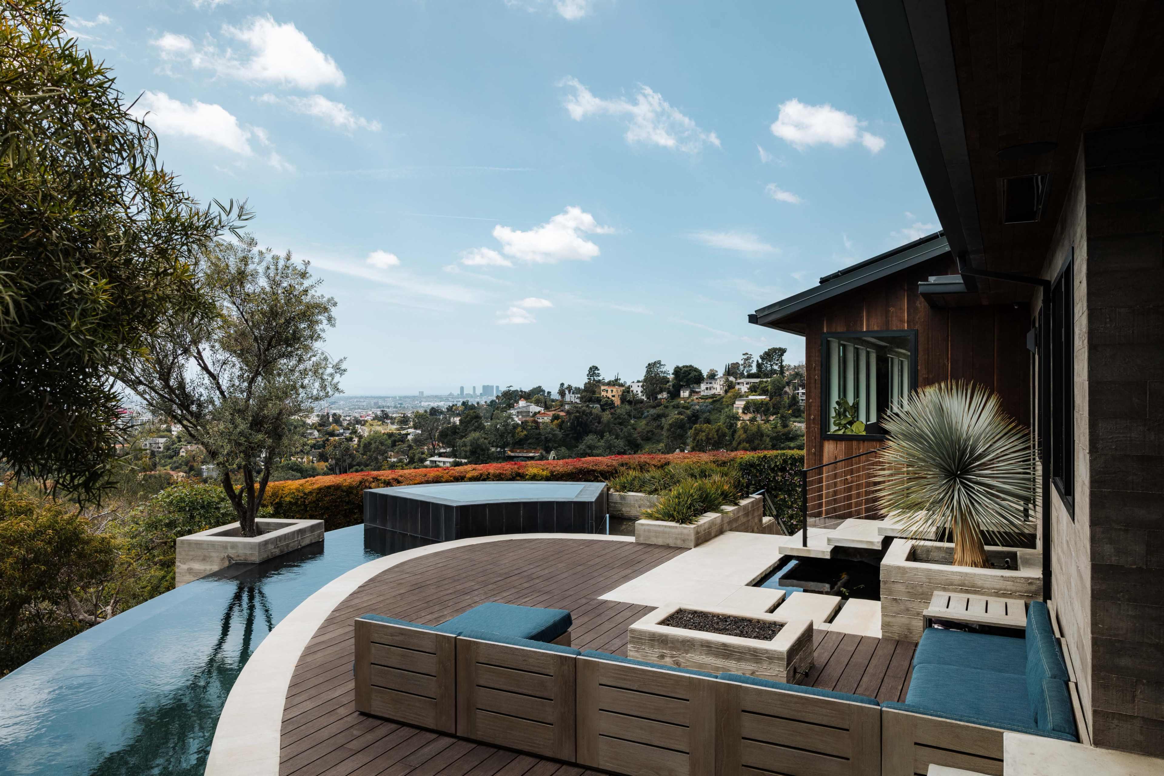 The image depicts a modern outdoor living space with a wooden deck, a small pool, and stylish seating, overlooking a scenic view of a city and surrounding hills.