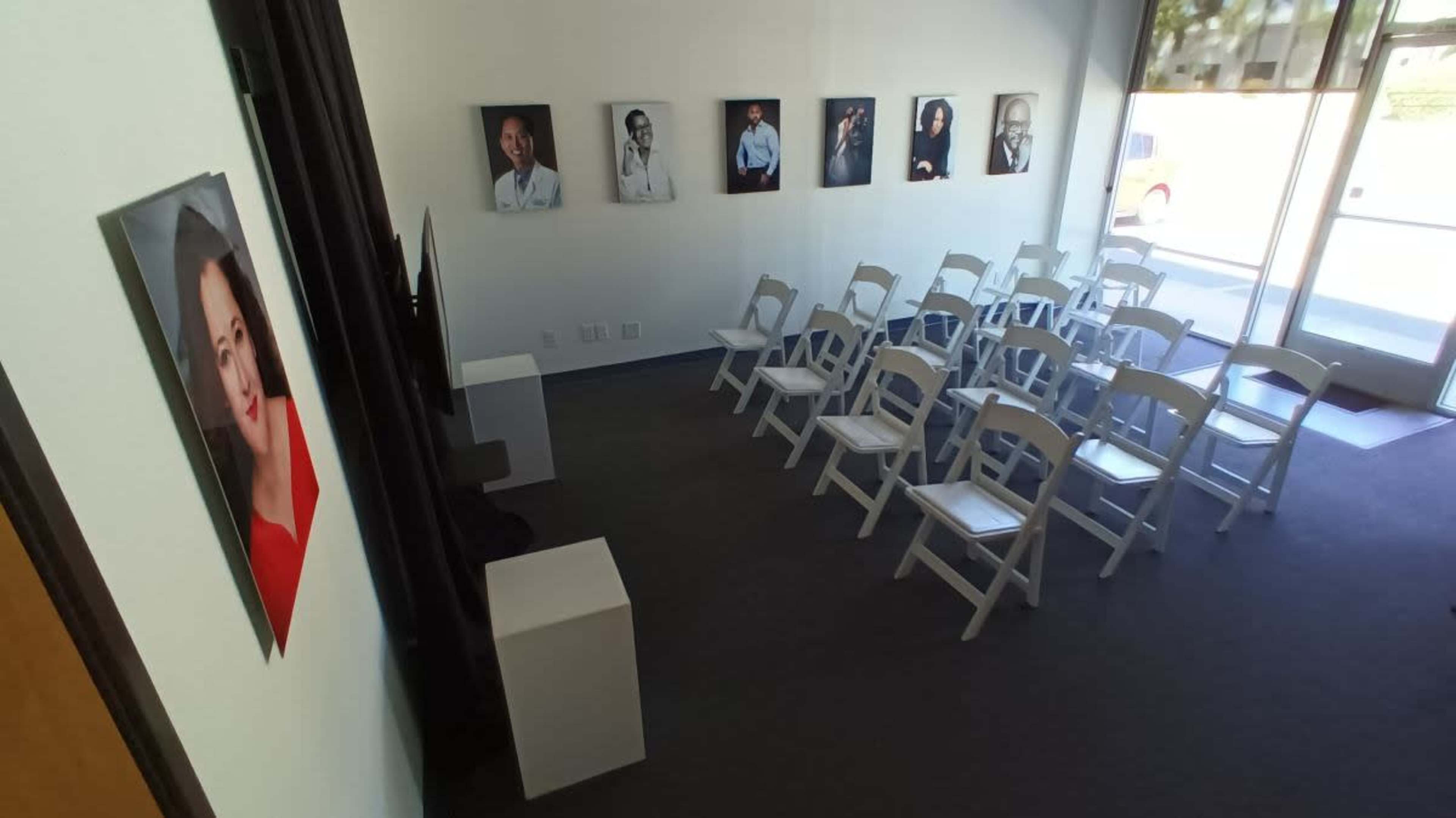 The image shows an interior room with several white folding chairs arranged in rows and framed portraits hanging on the walls.