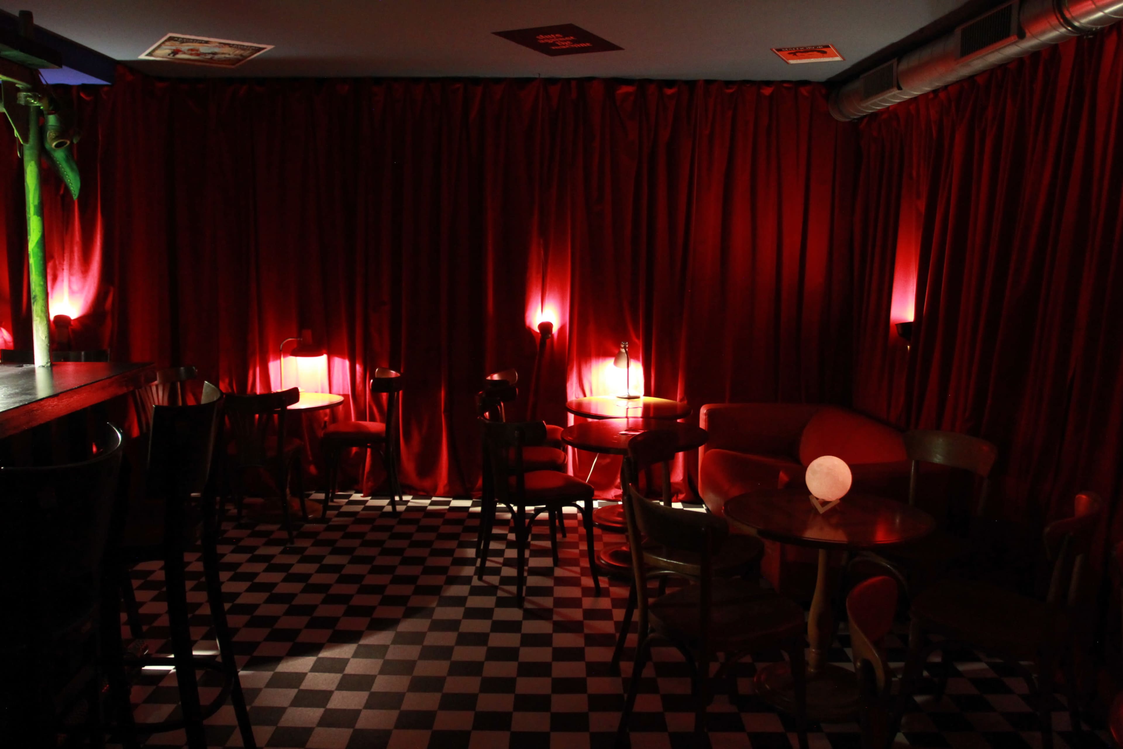 The interior of a dimly lit bar features red curtains, checkered flooring, and small tables illuminated by soft lighting.