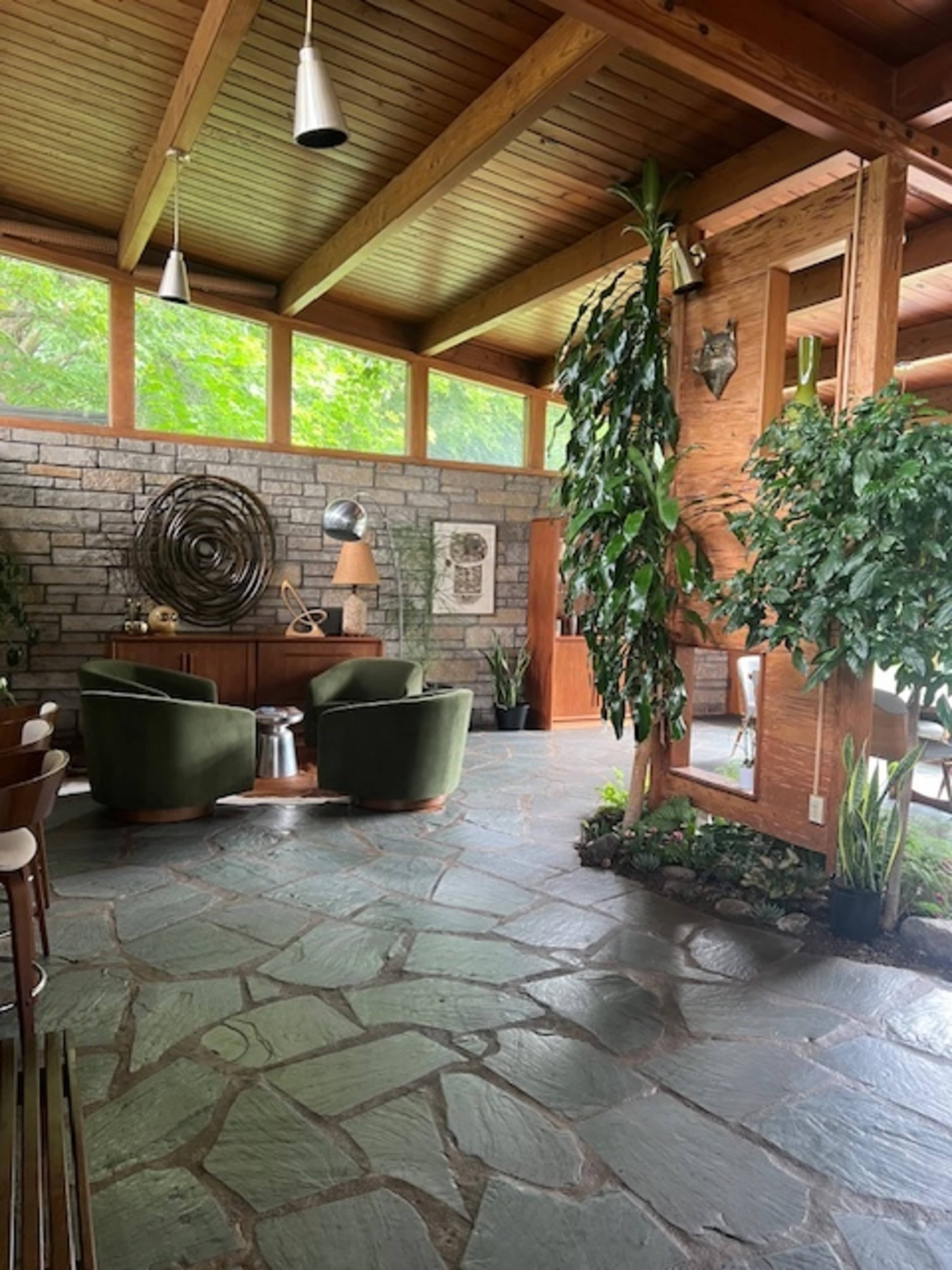The image shows a spacious interior featuring stone flooring, wooden beams, two green armchairs, and tropical plants.