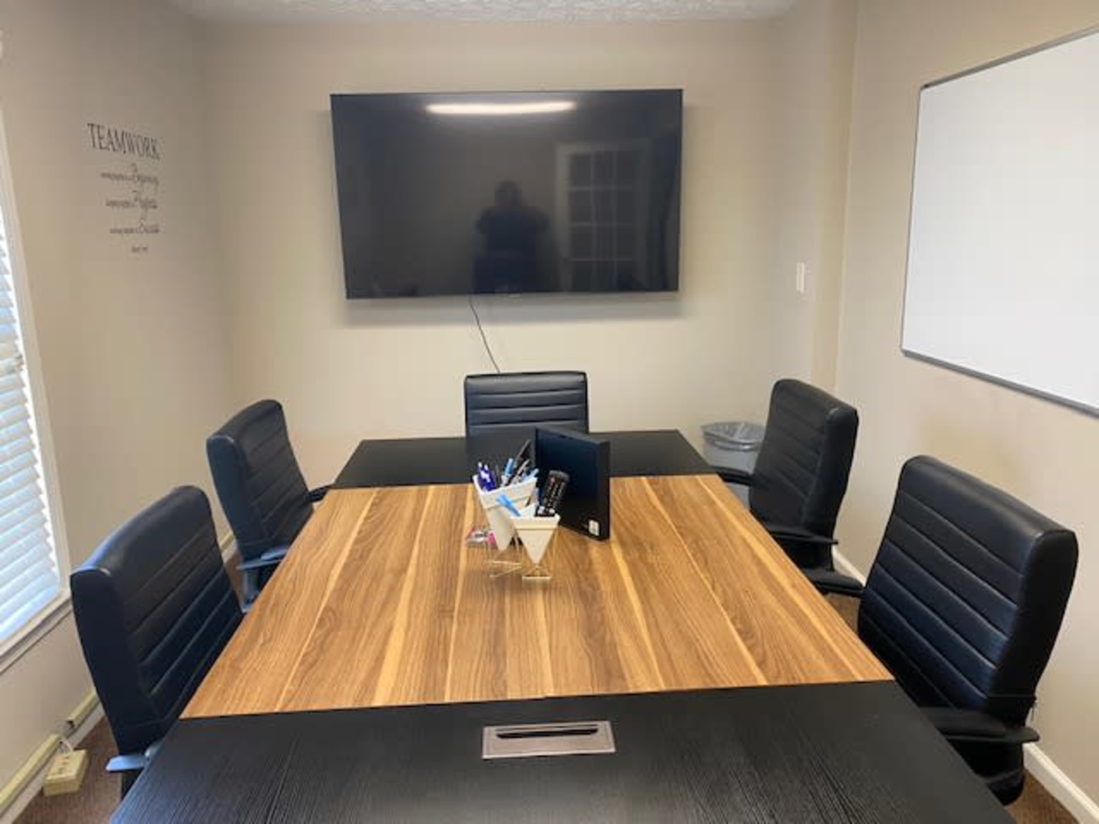 A conference room features a large wooden table surrounded by black chairs, a wall-mounted TV, and a whiteboard.