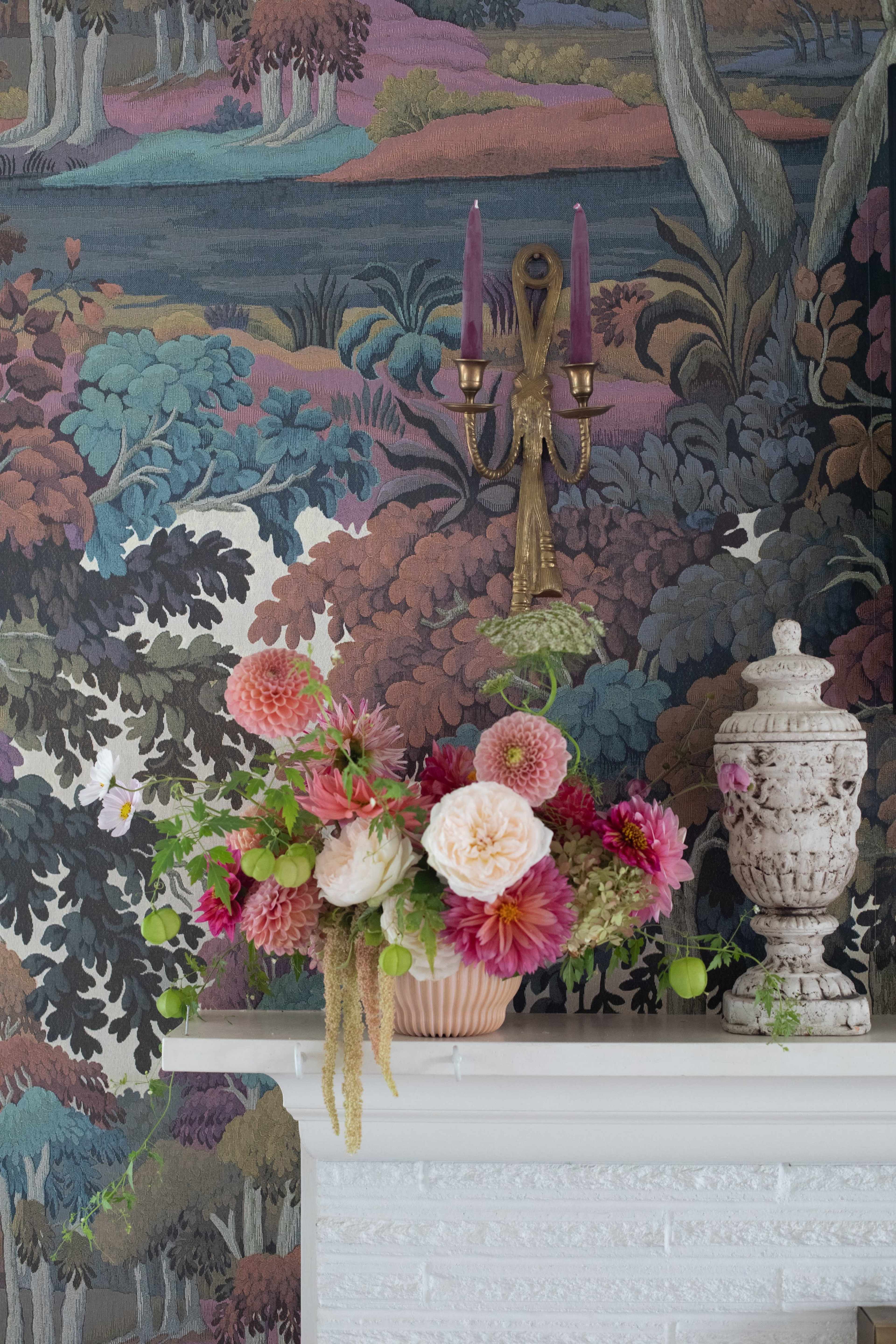 A floral arrangement with various blooms is placed on a white mantelpiece in front of a colorful, patterned wallpaper depicting a wooded landscape.
