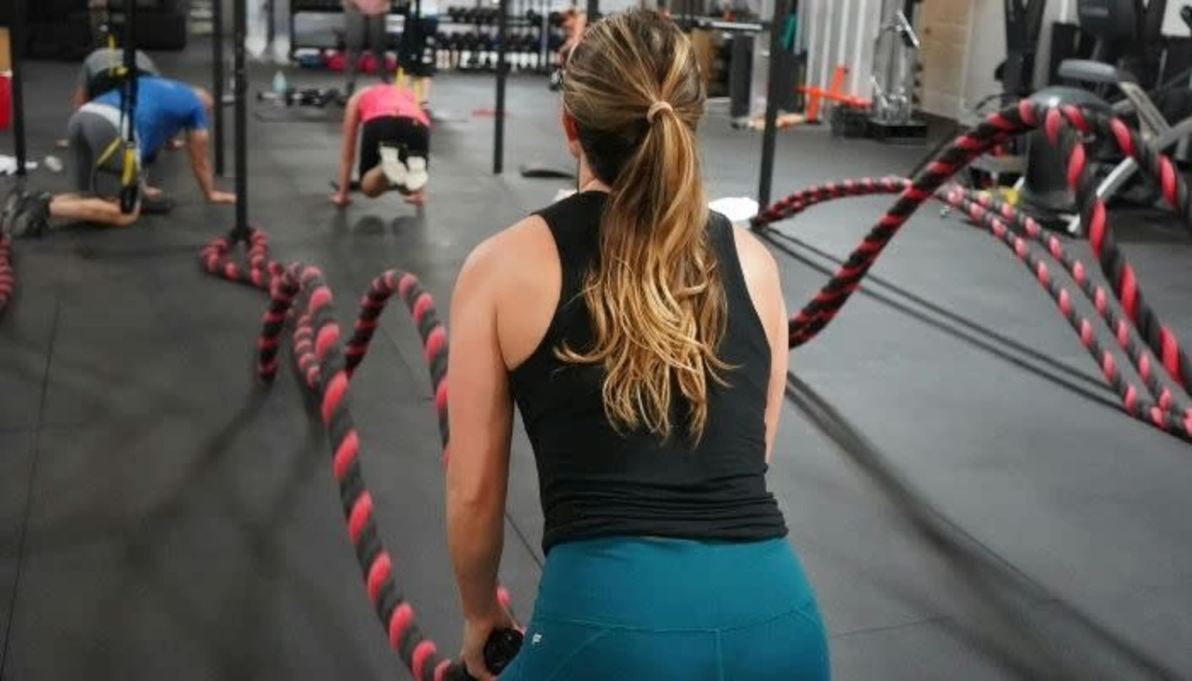 A woman exercises with battle ropes in a gym while others perform various workouts in the background.