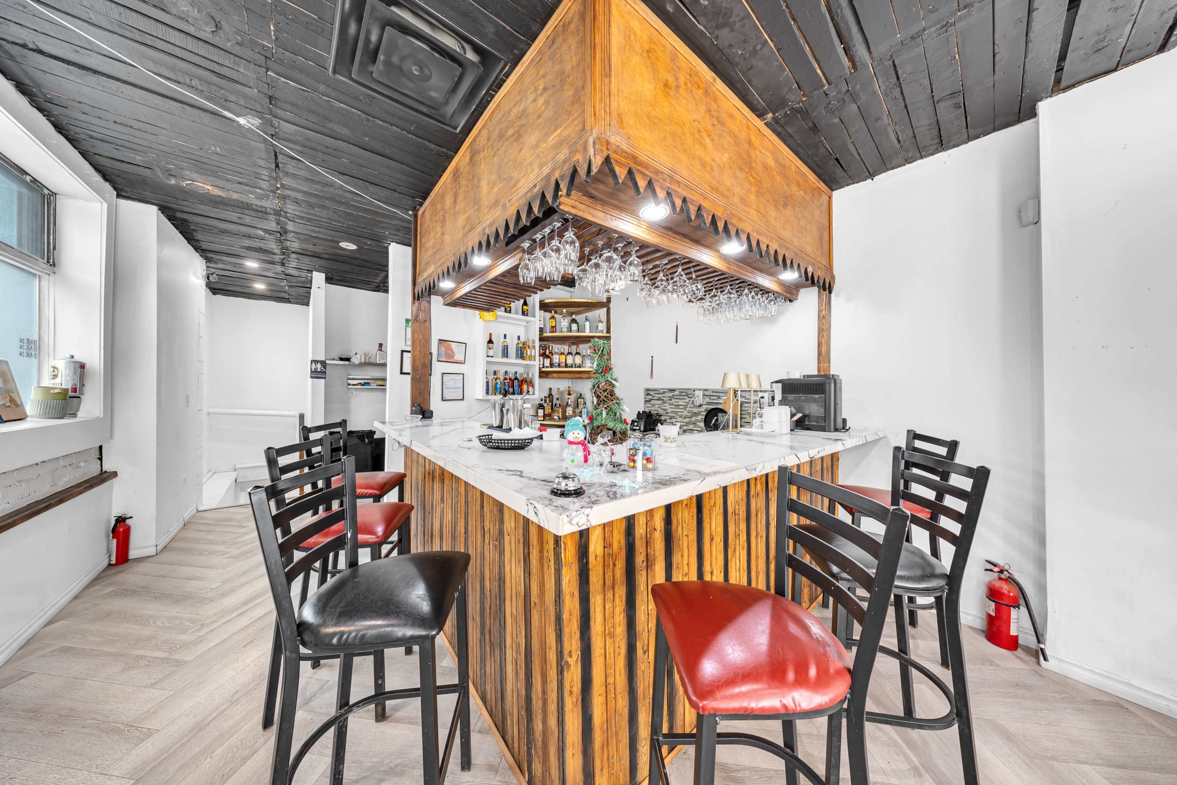 The image shows a modern bar area with a wooden counter, stools, and a selection of bottles displayed on shelves behind it.