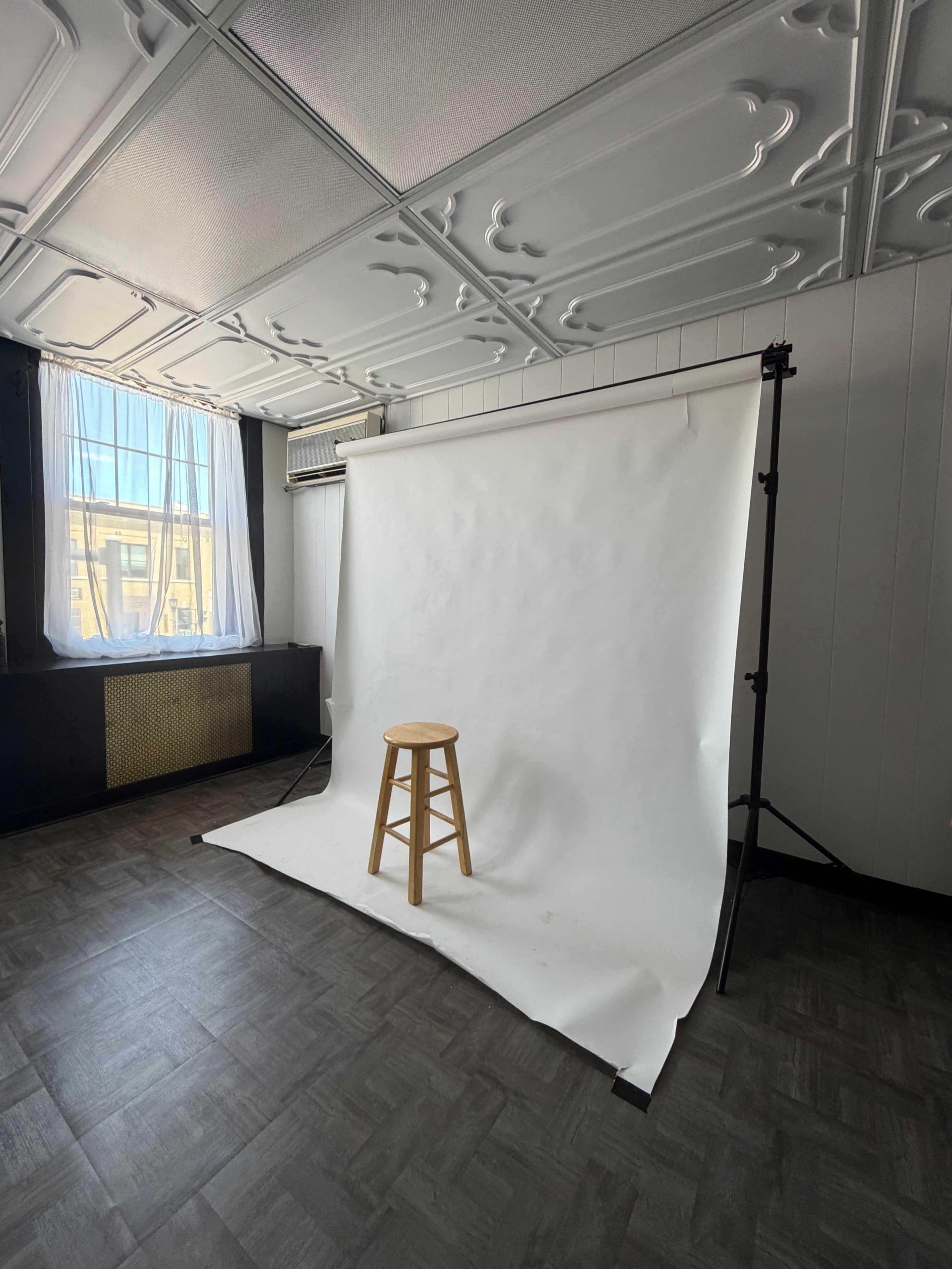 The image shows a minimalist interior space featuring a wooden stool in front of a plain white backdrop, with natural light coming through a window.